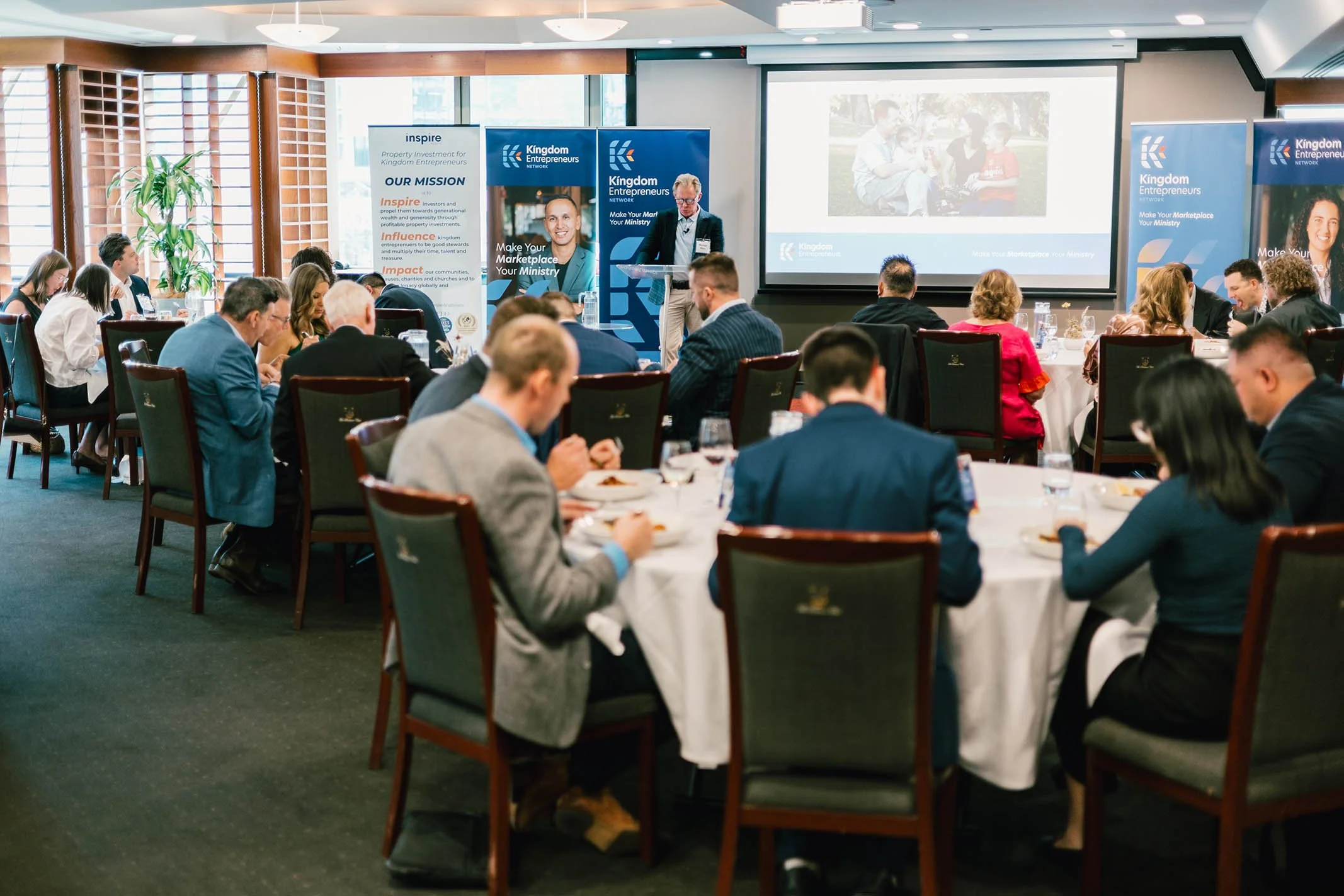 Business conference in a room with round tables, attendees listening to a speaker at a podium, slide projections, and promotional banners for Kingdom Entrepreneurs Network.