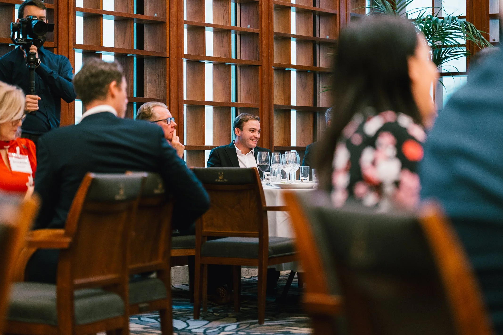 People seated at a table during a formal event or conference, with a man smiling and wearing a suit, and others engaged or listening, in a room with wooden wall paneling and large windows.