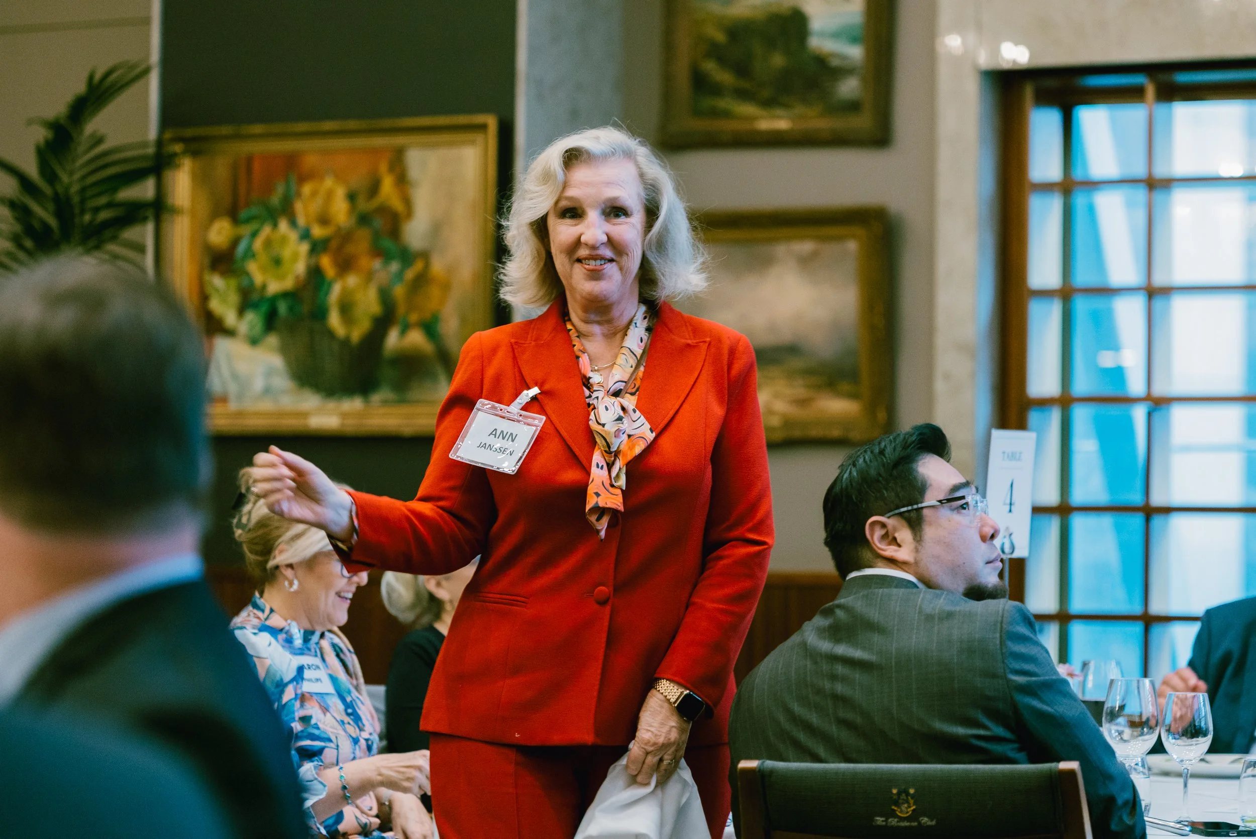 A woman with blonde hair, wearing a red blazer and colorful scarf, standing and smiling at a formal event with seated guests at a table.