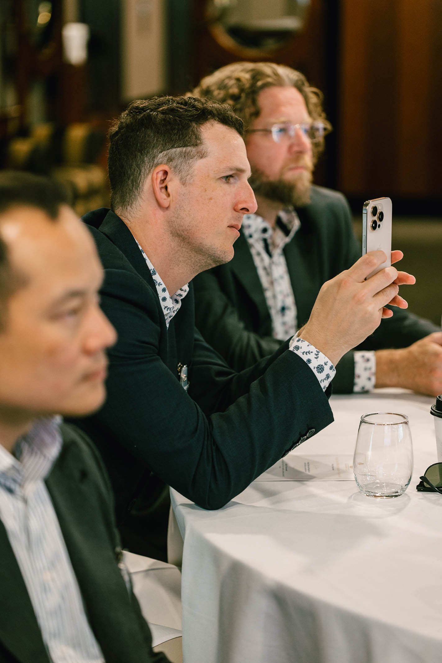 A man in a dark suit and white shirt with floral pattern, sitting at a white table with a glass of water, is focused on something off-camera. Behind him, another man with curly hair, glasses, and a patterned shirt under a dark blazer, is holding a sm