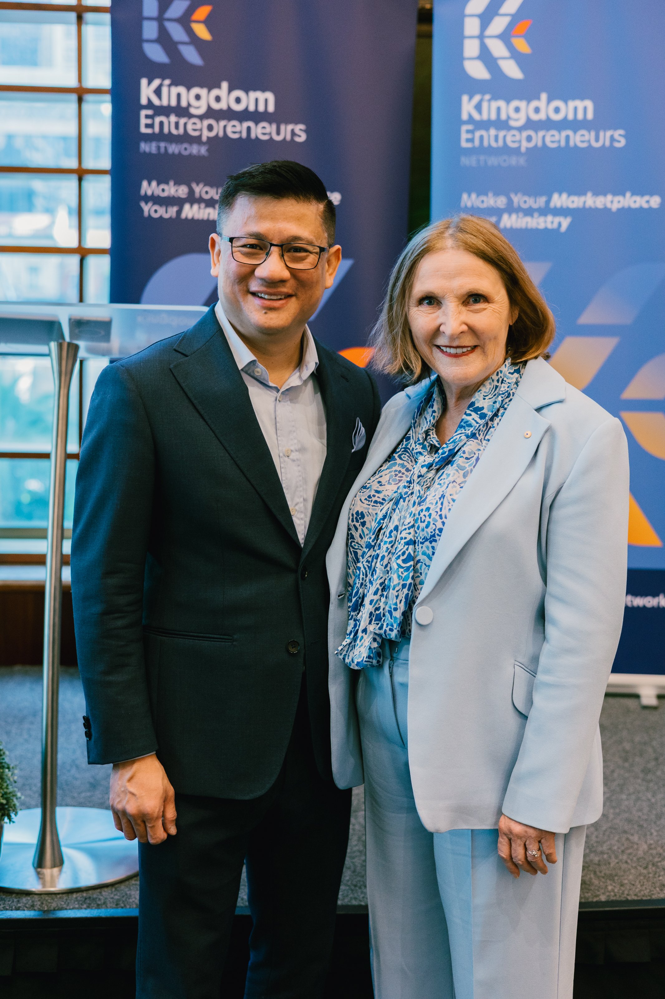 Two people, a man and a woman, standing together in front of a Kingdom Entrepreneurs banner at an event, smiling at the camera.
