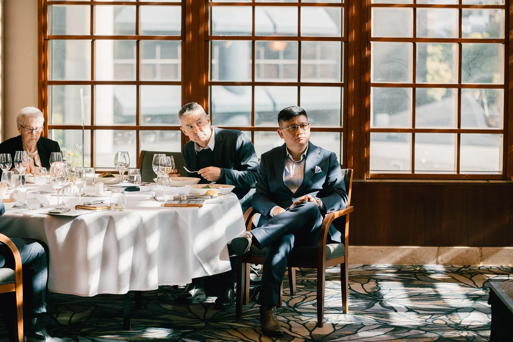 People attending a formal meeting or conference in a well-lit room with large windows, some looking attentive and engaged.
