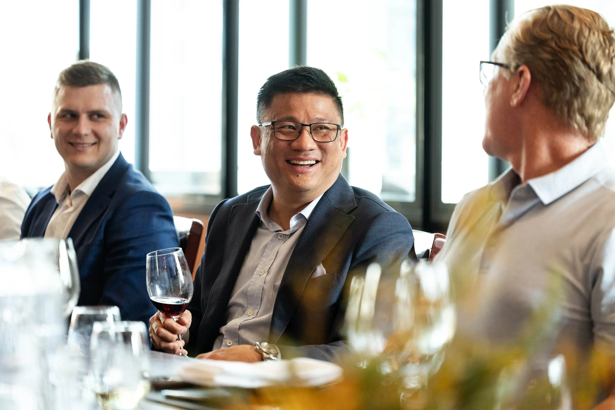 Three men in business attire sitting at a table, smiling and engaging in conversation during a social or professional gathering, with wine glasses and table settings in front of them.