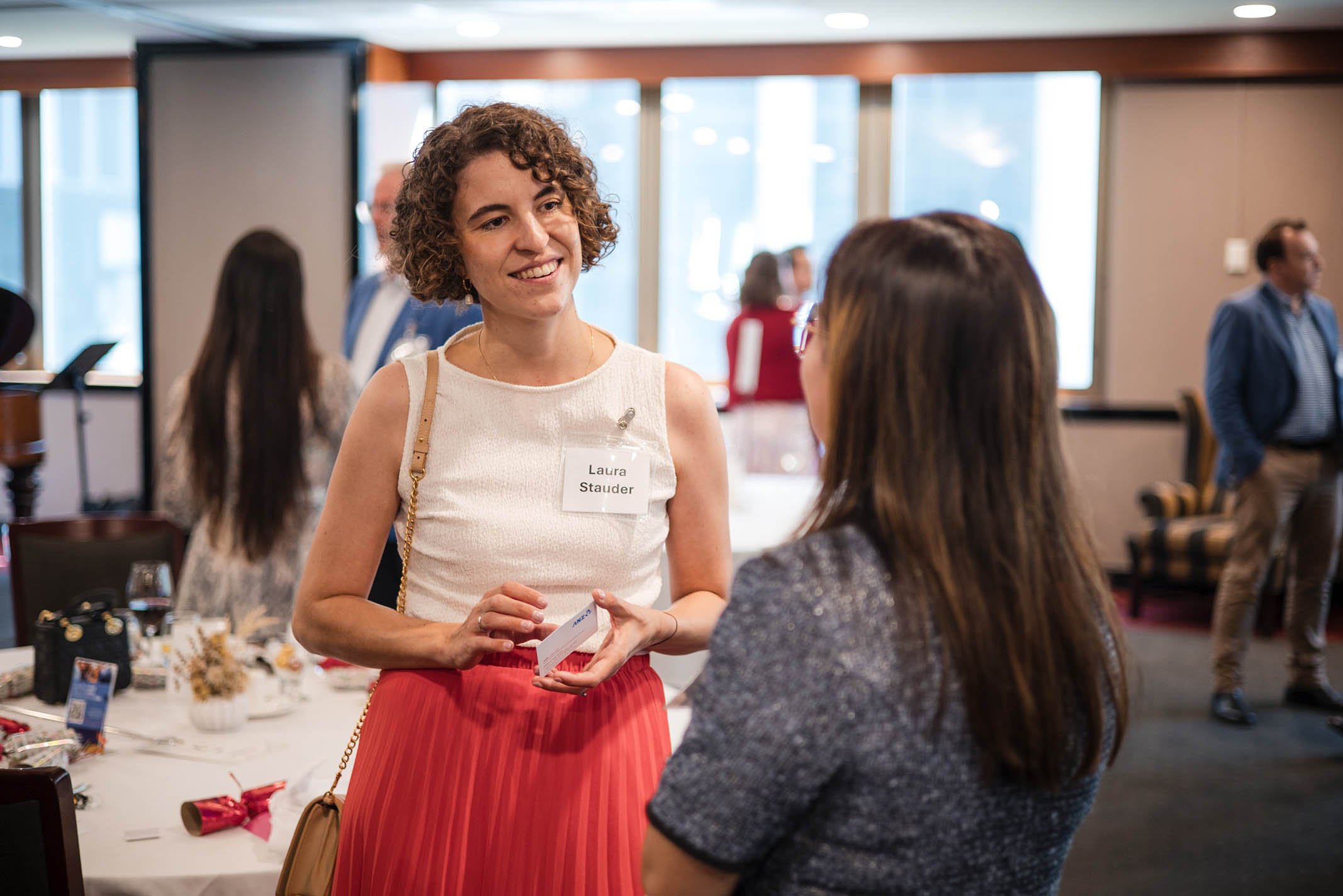 Two women engaged in conversation at a networking event in a well-lit room with large windows, with other attendees visible in the background.