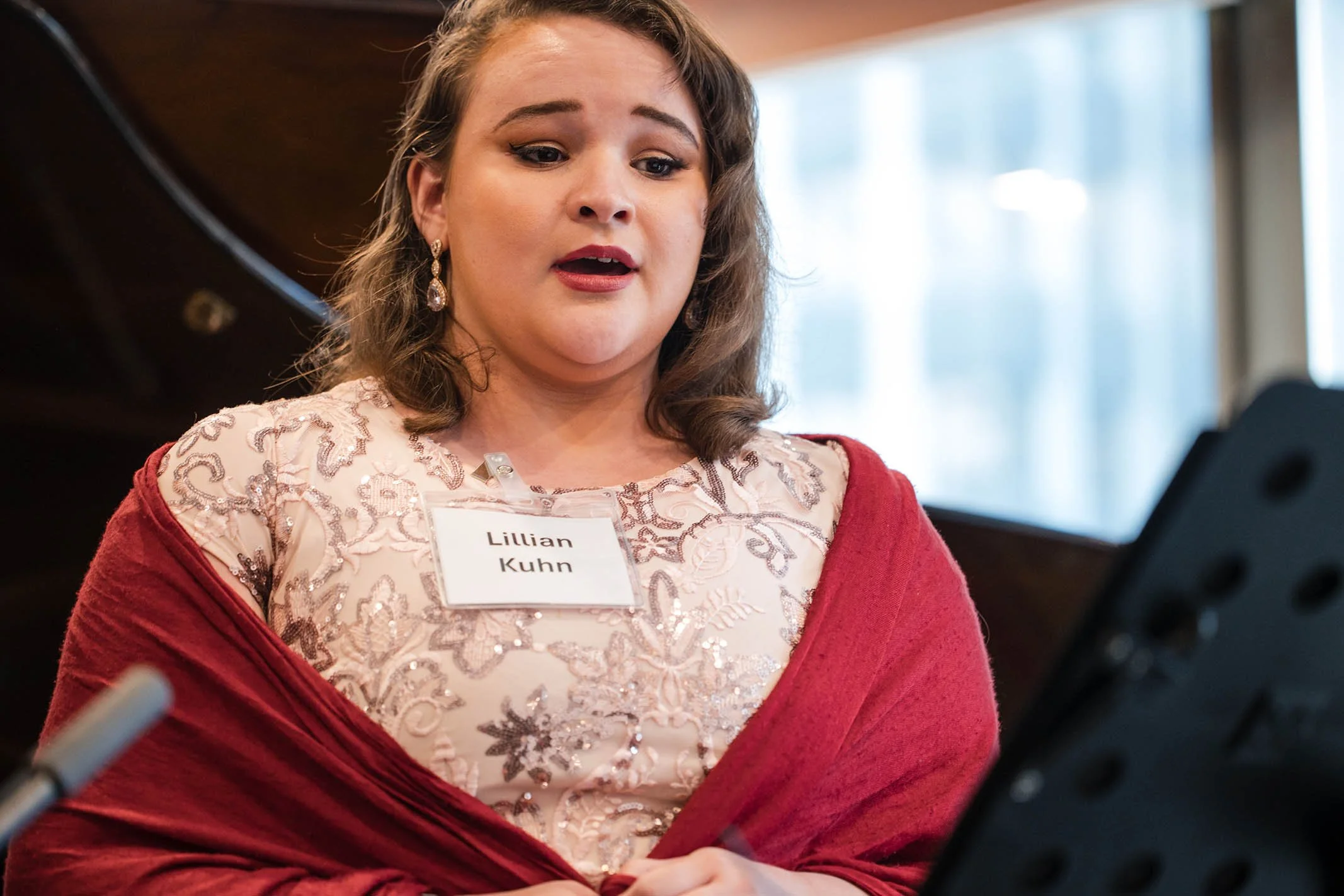 A woman wearing a pink embroidered dress with a red shawl, holding a sheet of music, looking focused, with a name tag that reads 'Lillian Kuhn', in a room with large windows.