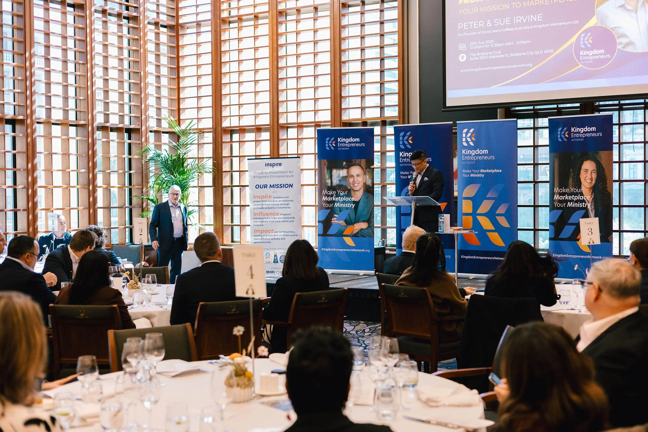A conference room with attendees seated at round tables, listening to a speaker at a podium. The room has large wooden window panels, a large digital screen, and banners displaying the 'Kingdom Entrepreneurs Network' logo and messages. The speaker is