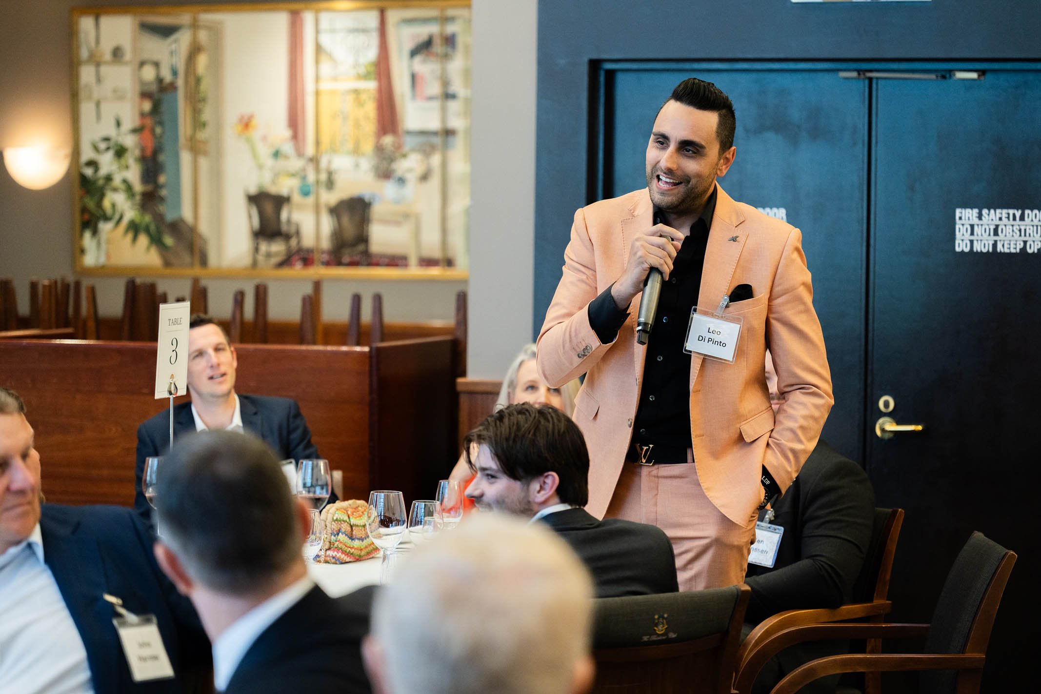 A man in a peach-colored suit standing and speaking into a microphone at a formal event. Seated around him are other professionally dressed people, some with name tags. The setting appears to be a banquet or conference room.