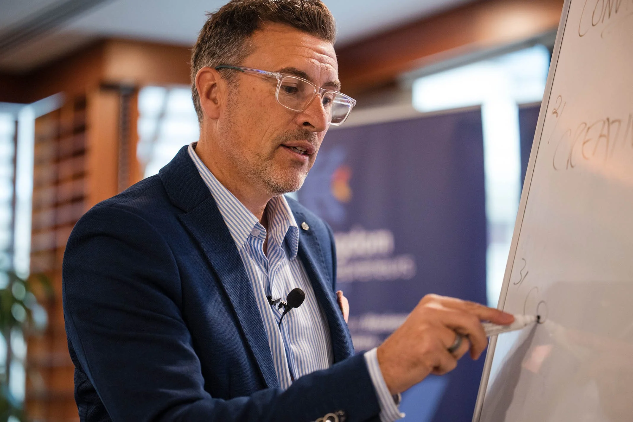 A man in a dark blue blazer, striped shirt, and glasses points at a whiteboard during a presentation.