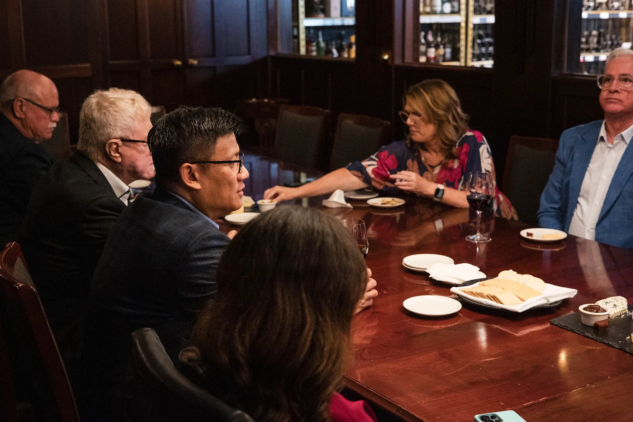 A group of people sitting around a large wooden table in a upscale restaurant or private dining room. They are engaged in conversation, with some eating and drinking. The table has glasses of wine and plates with snacks and desserts.
