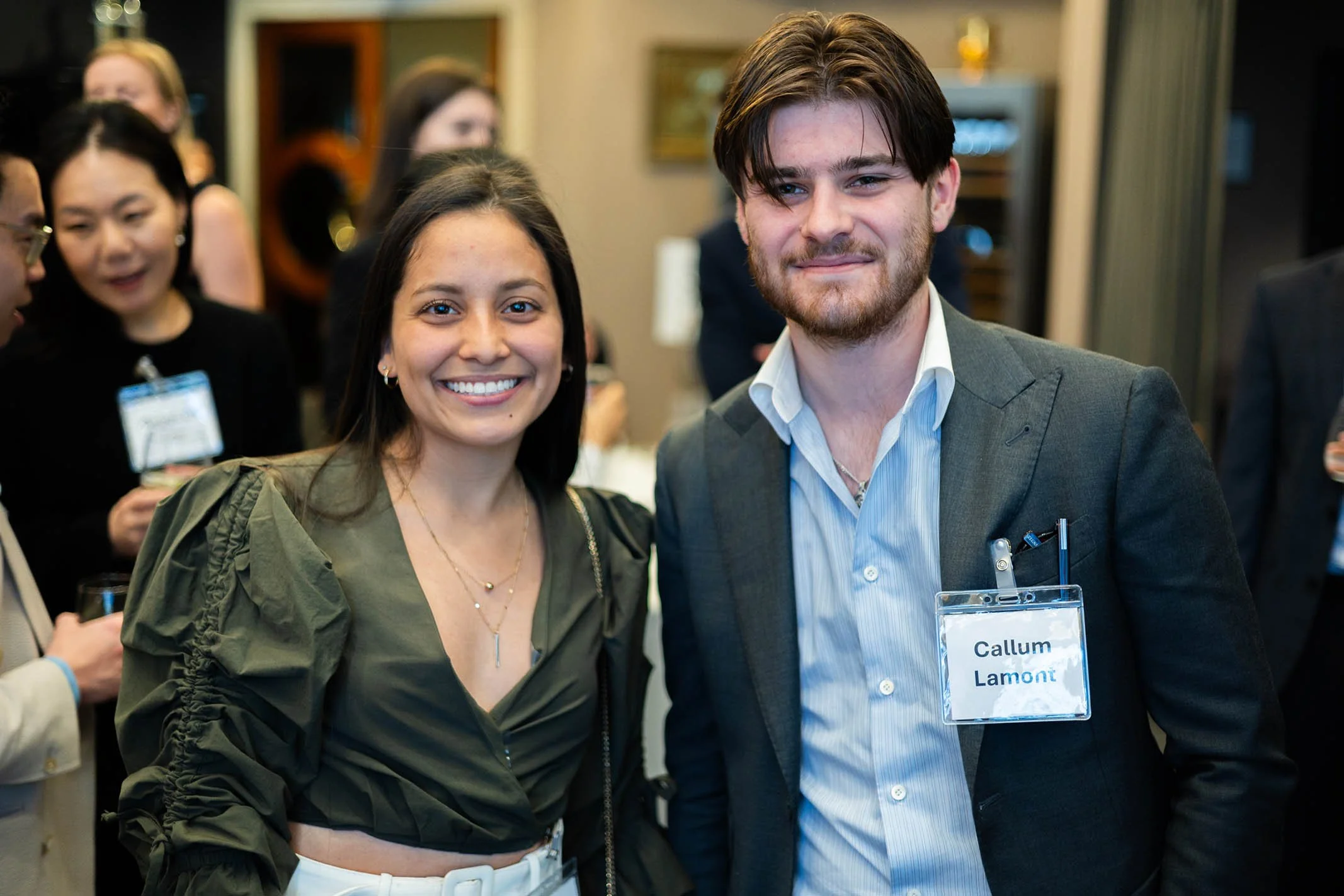 A woman and a man posing together at a professional event, with other attendees in the background.