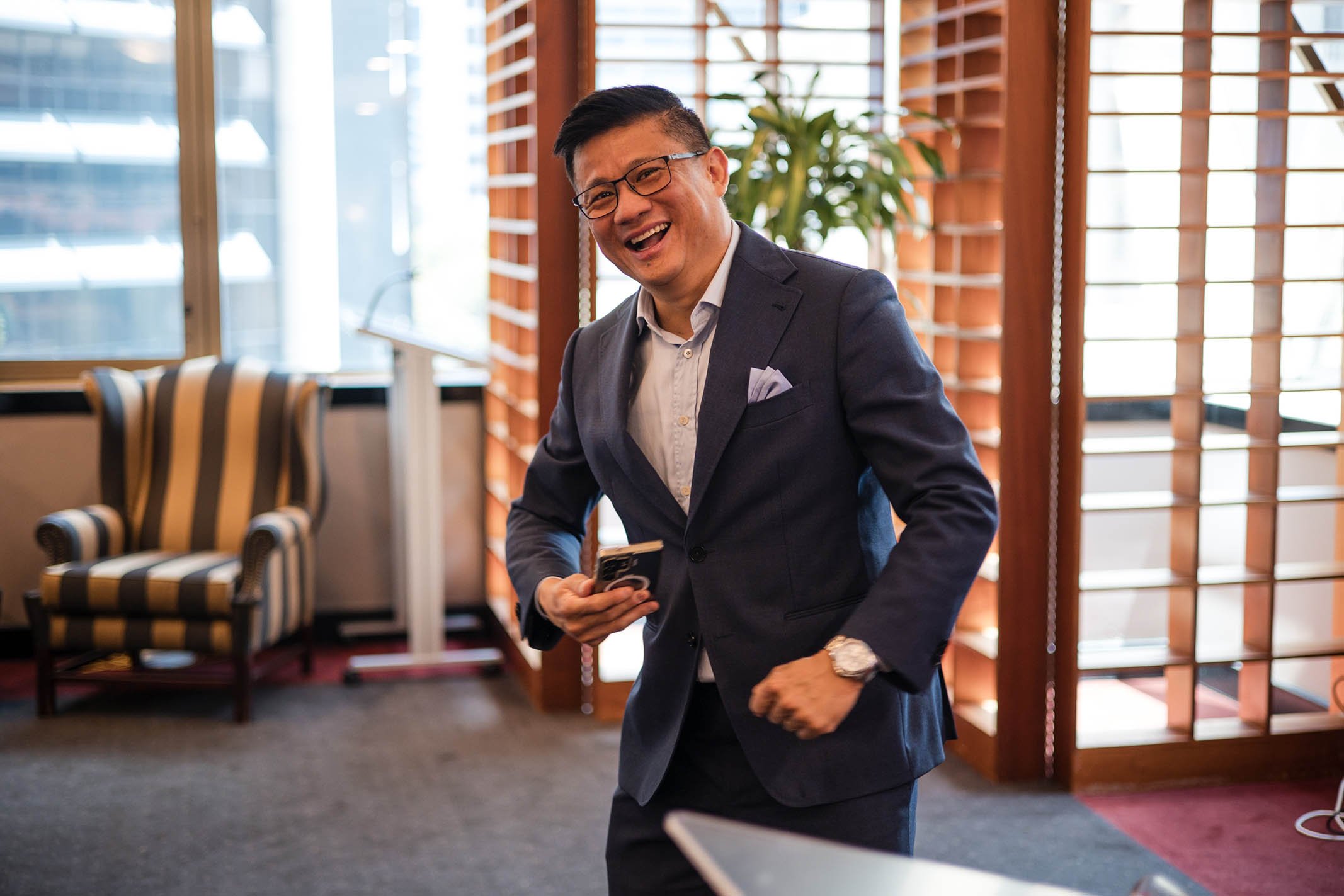 A man in a dark suit and glasses, smiling and laughing, holding a smartphone in a well-lit indoor space with wooden window shutters, a striped armchair, and houseplants.