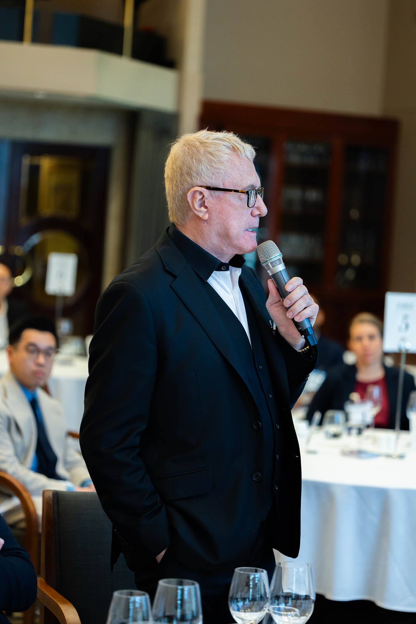 A man with blond hair, glasses, and a black suit is speaking into a microphone at a formal event. People are seated at tables in the background.