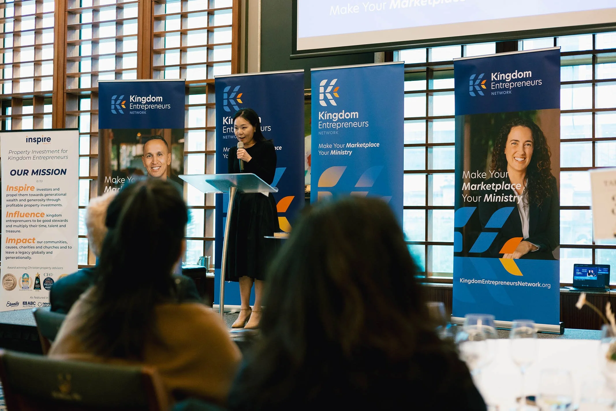 A woman giving a speech on a stage at a conference with banners behind her displaying 'Kingdom Entrepreneurs Network' and portraits of other speakers. The audience is seated, listening to the presentation.