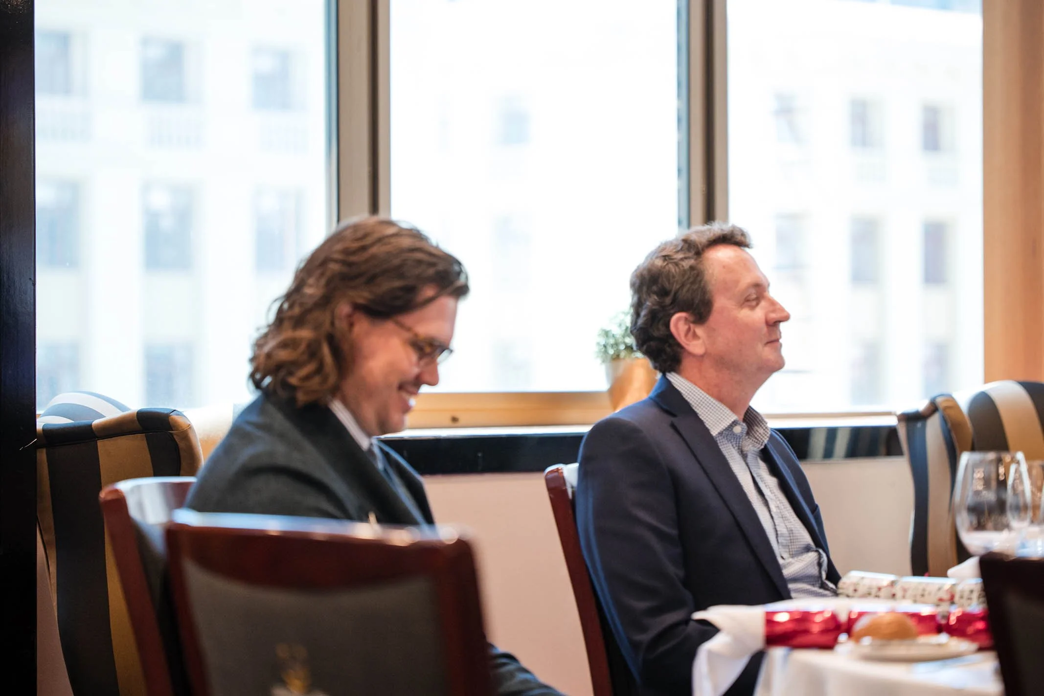 Two men in suits sitting at a dining table in a restaurant or conference room, smiling and appearing relaxed. A window with a cityscape backdrop is visible behind them.