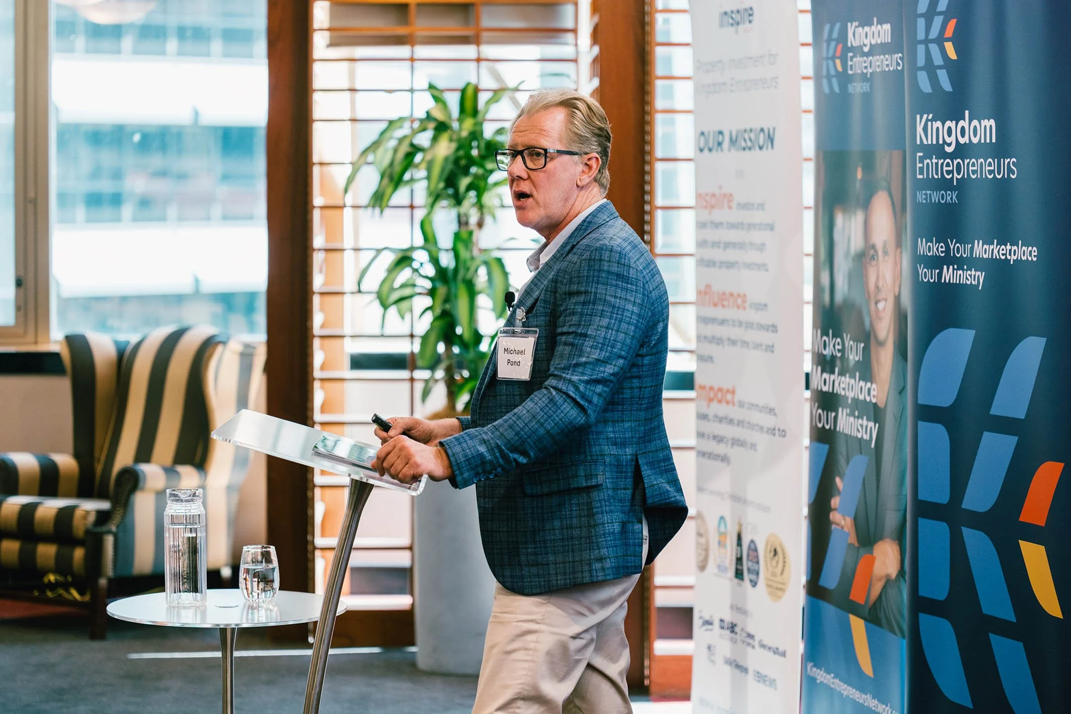 A man with glasses and gray hair, wearing a blue checkered blazer, white shirt, beige pants, and a name badge, speaking at a conference. He is standing behind a small podium with a microphone and holding a remote. Behind him are banners displaying th