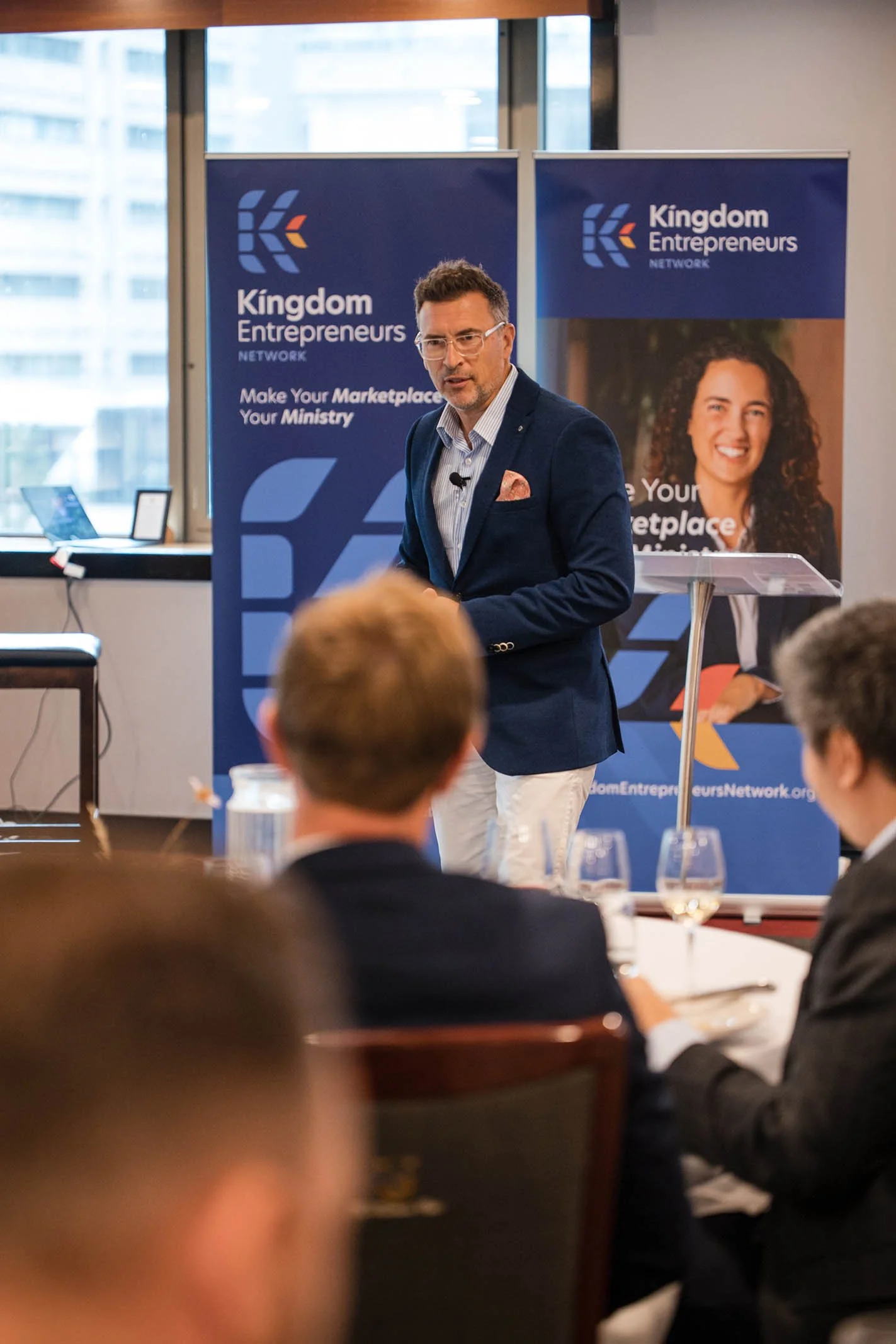 A man in a navy blazer and white pants giving a presentation at a conference, standing in front of banners with 'Kingdom Entrepreneurs Network' logo and slogan, with seated audience members watching.