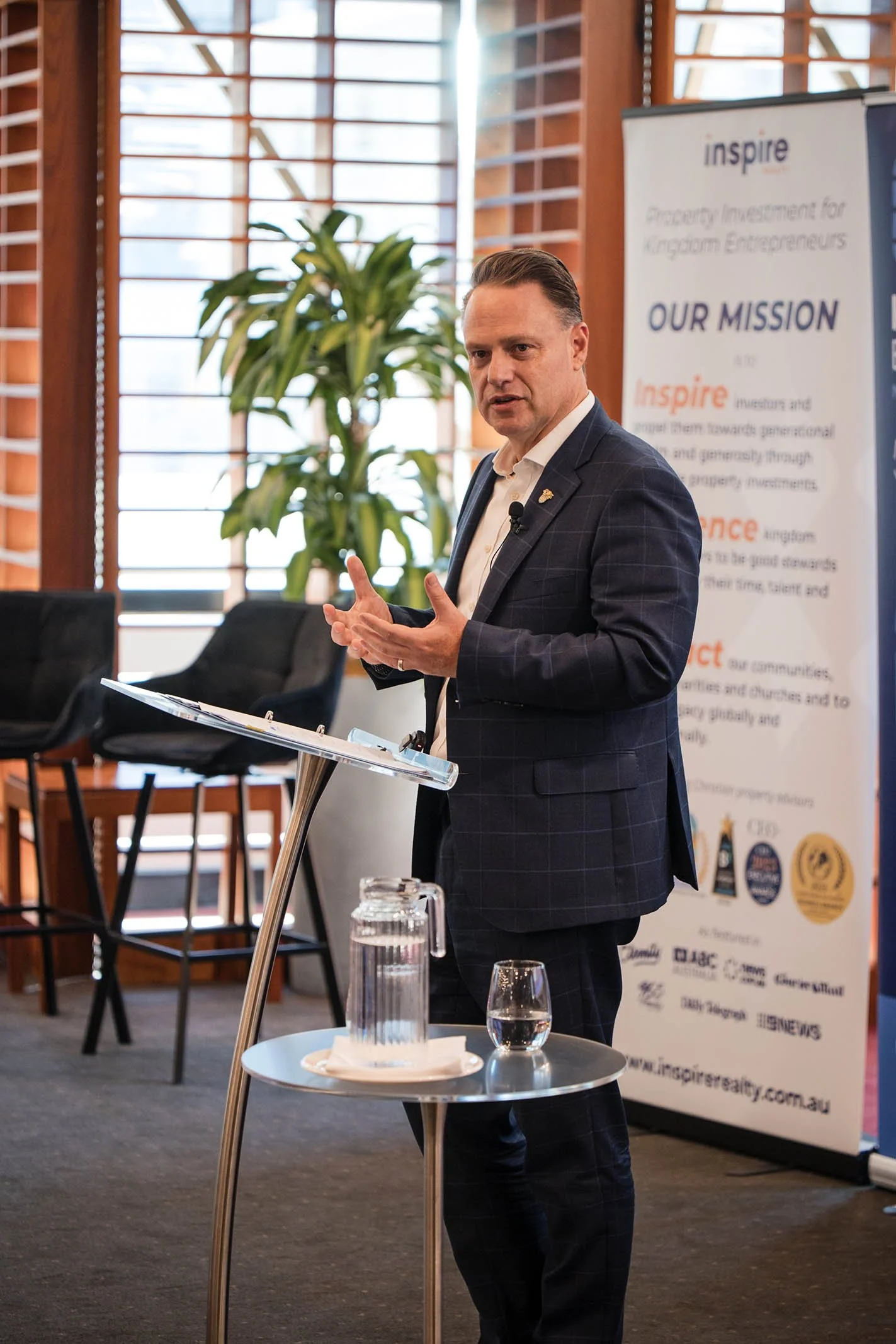 A man in a dark suit with a white shirt giving a presentation at a conference, standing next to a small round table with a glass of water and a pitcher. Behind him is a vertical banner with text and logos, and large windows with wooden blinds.