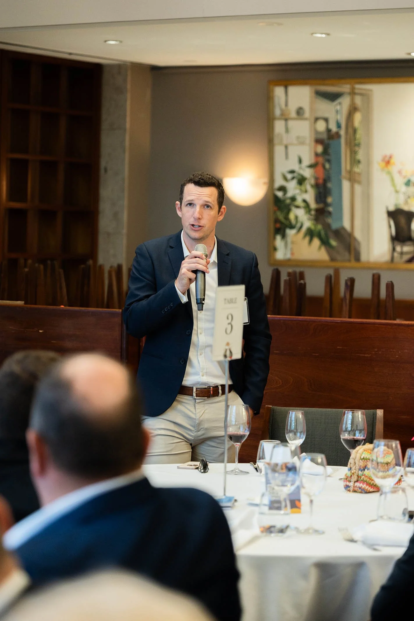 A man in a dark blazer and white shirt speaking into a microphone at a formal dining event, with several wine glasses and a colorful bag on the table, and a framed painting in the background.