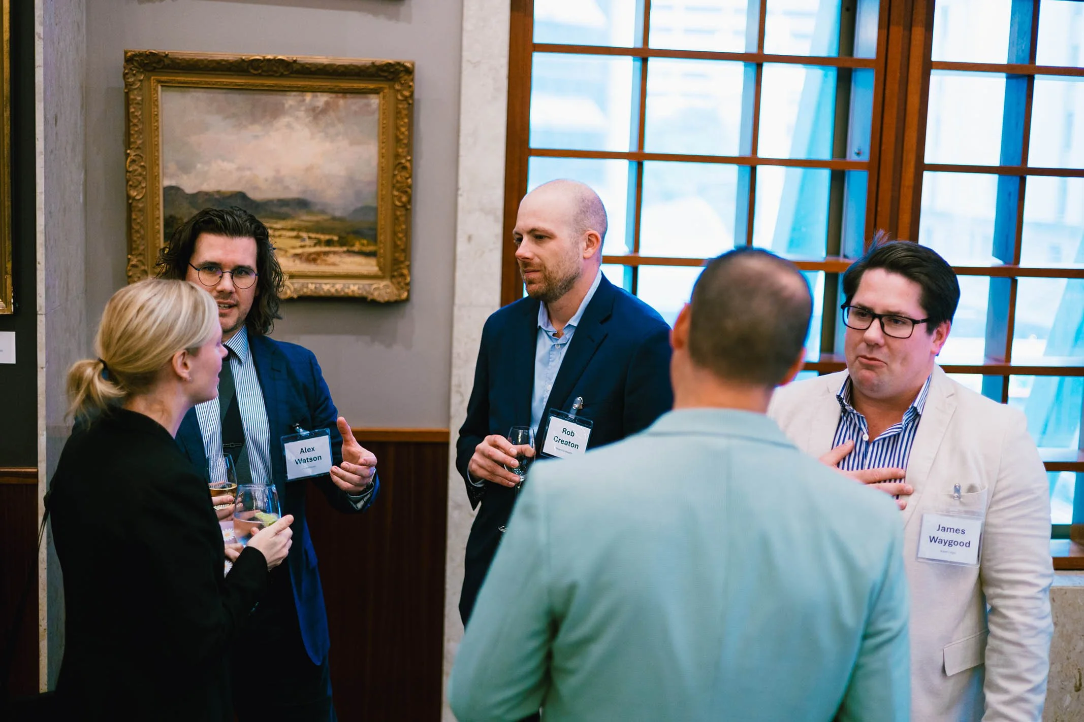 Group of five professionals having a conversation at a networking event, including two men and a woman with glasses, with paintings and large windows in the background.