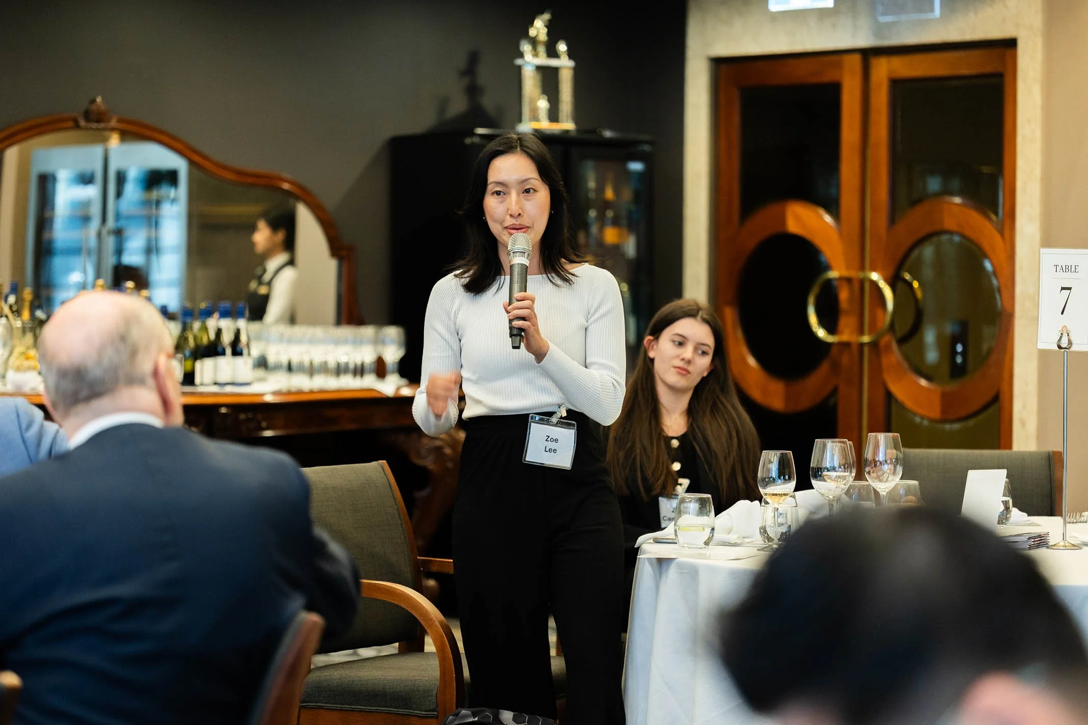 A woman holding a microphone speaking to a group of people at a formal event or conference in a restaurant or banquet hall setting.