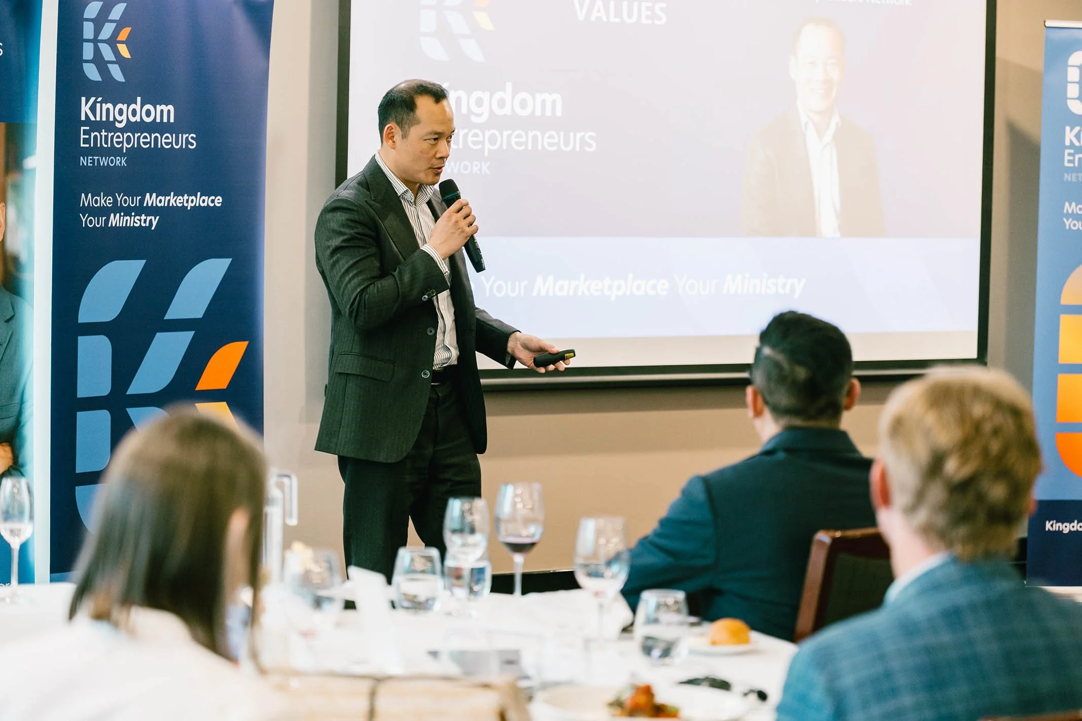 A man in a dark suit giving a presentation at a conference, holding a microphone and a remote. Audience members are seated at tables with glasses of water, facing him. Behind him is a screen displaying the text 'Kingdom Entrepreneurs' and the slogan 