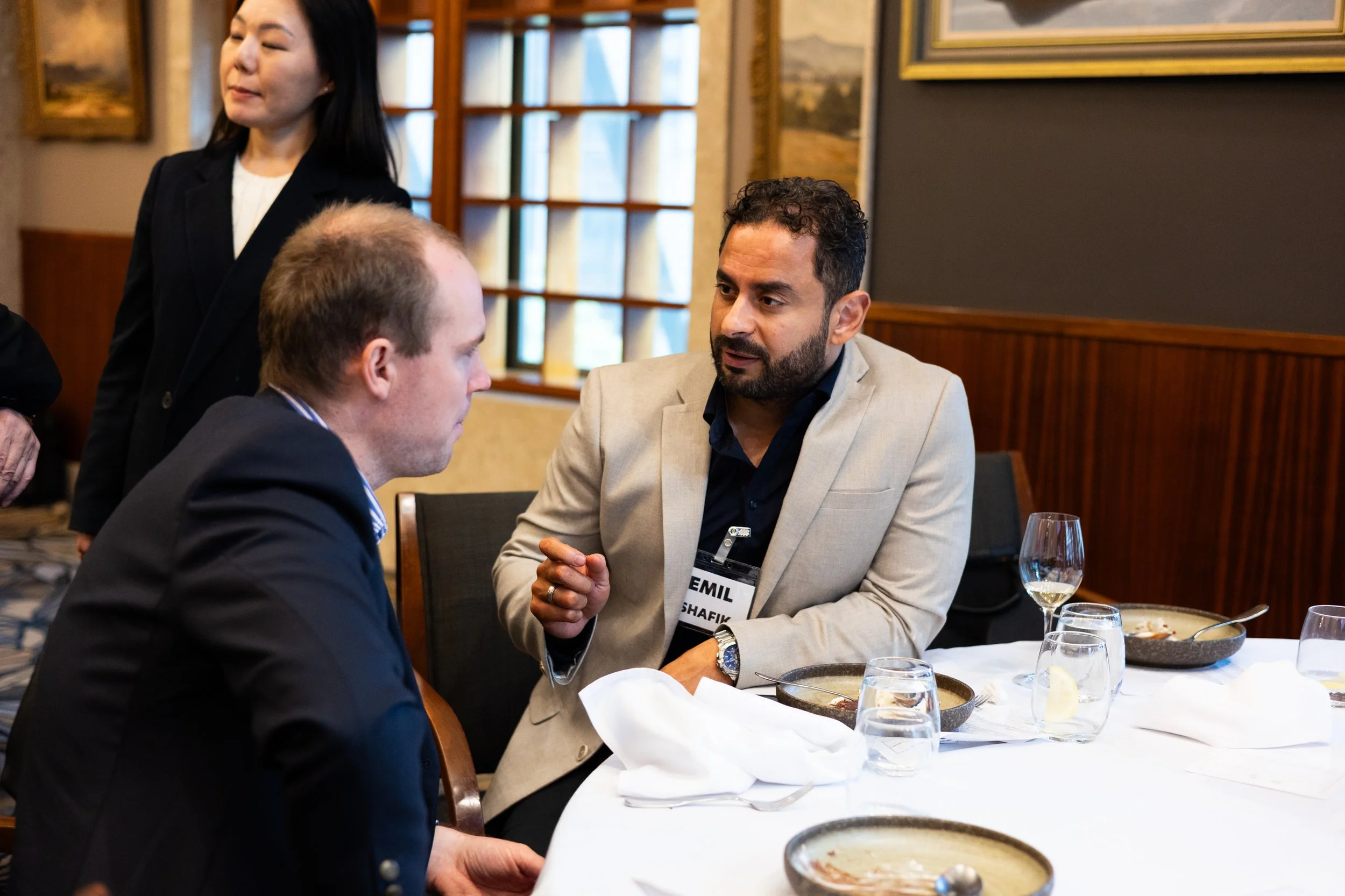 Two men engaged in conversation at a dining table during a professional event, with a woman standing nearby, in a restaurant or conference room setting.