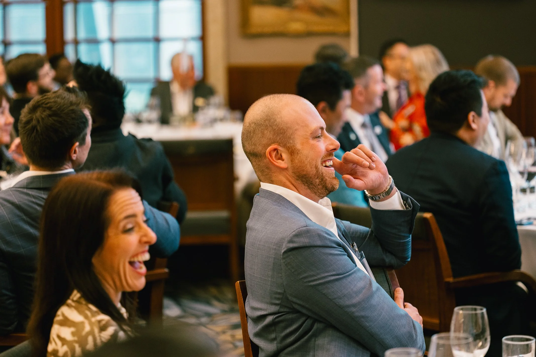 A group of professionally dressed people sitting at a banquet table, smiling and laughing during an indoor formal event or conference.