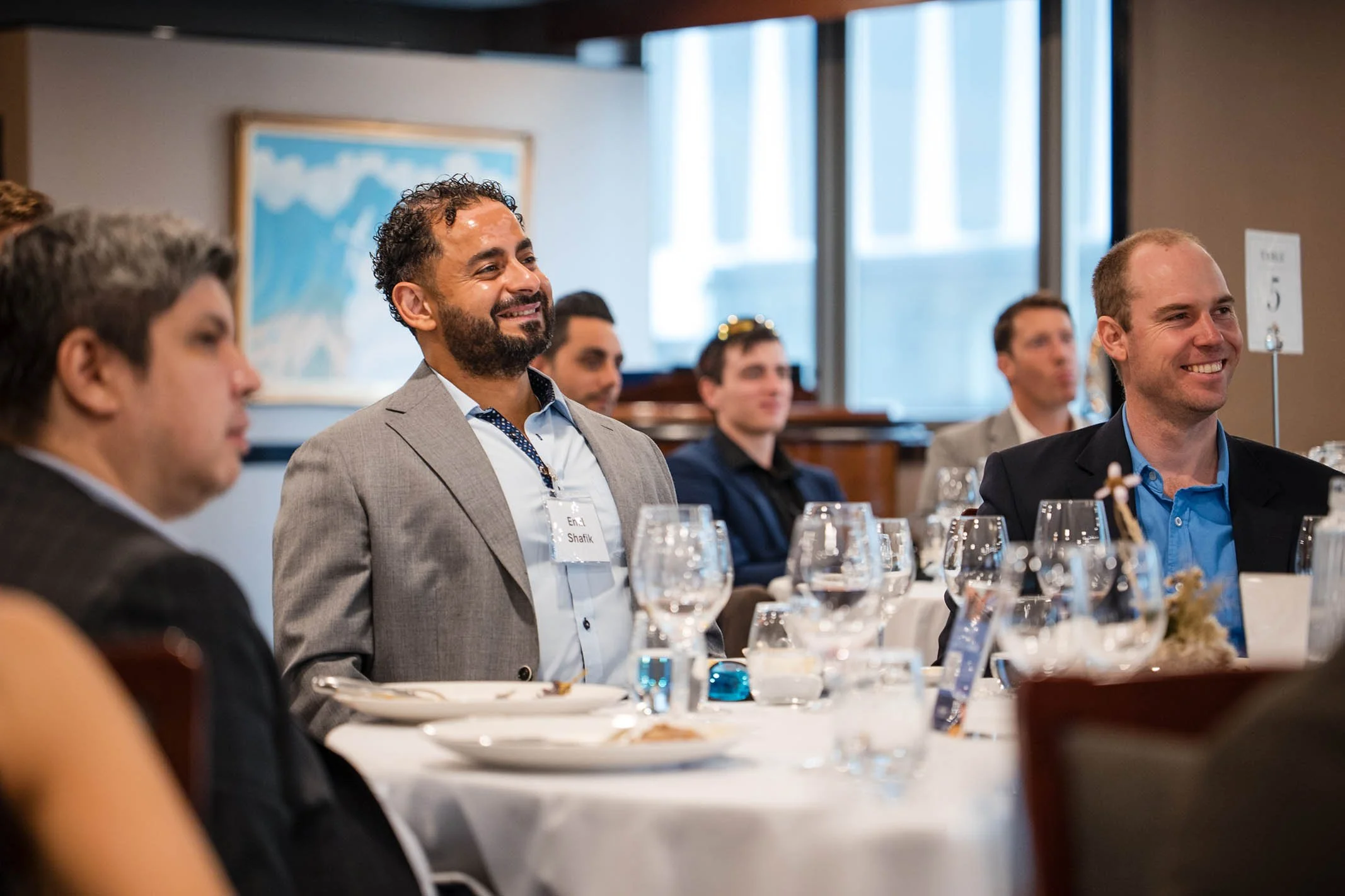 People attending a formal event or conference, seated at tables with white tablecloths, water glasses, and menus, smiling and listening to a speaker.