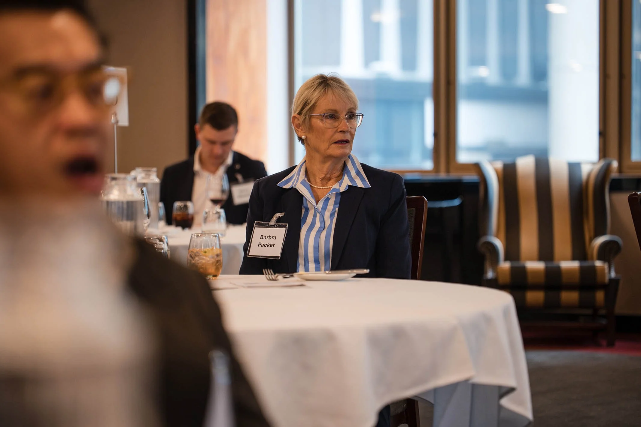 A woman with short blonde hair, glasses, and a blue striped shirt under a dark blazer, sitting at a round table during a conference or meeting. She has a name tag that reads 'Barbra Packer'. Other attendees are visible in the background.