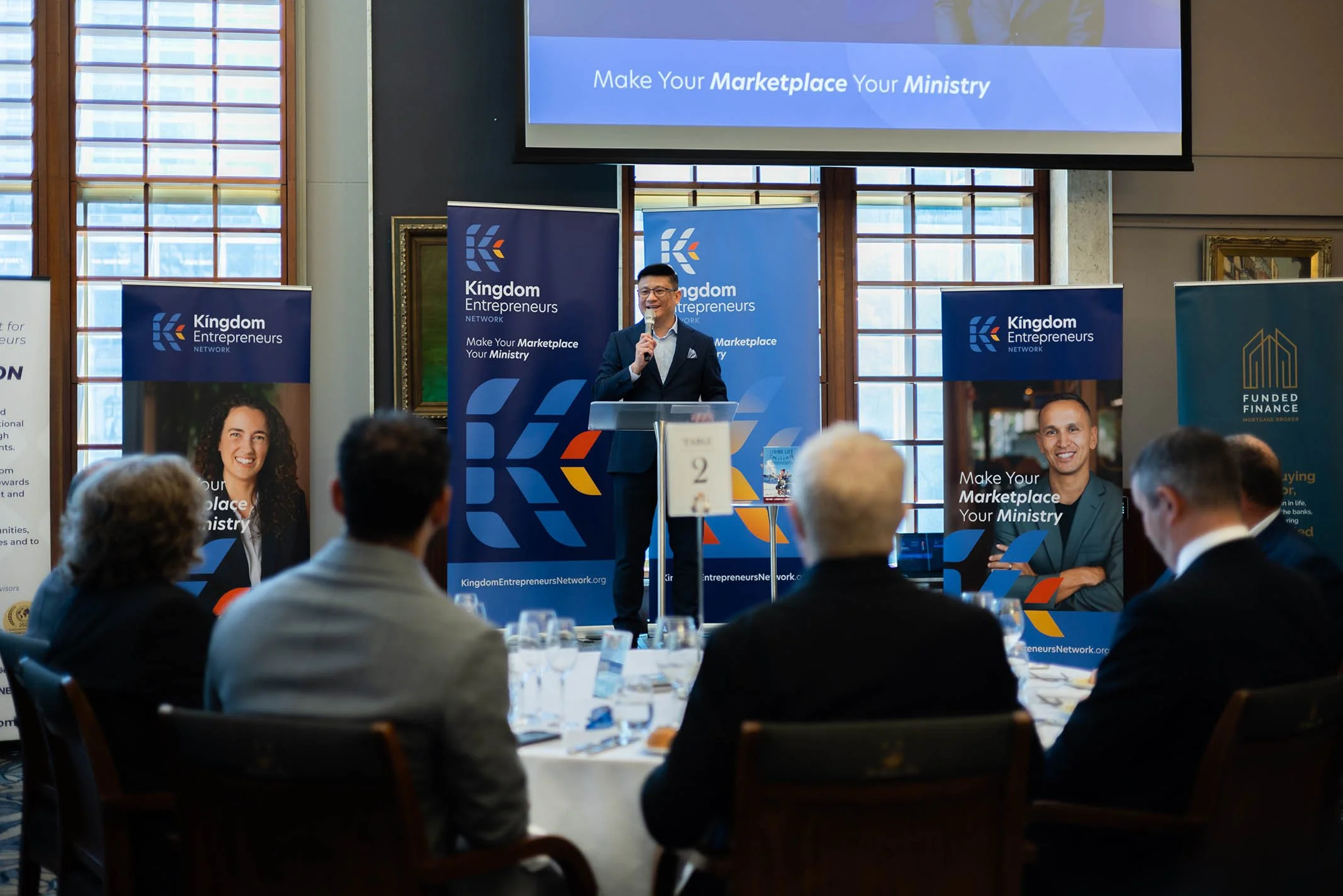 A man in a suit standing at a podium speaking to an audience at a conference or seminar. The event is related to 'Kingdom Entrepreneurs' and includes banners with the phrase 'Make Your Marketplace Your Ministry'. The room has large windows and attend