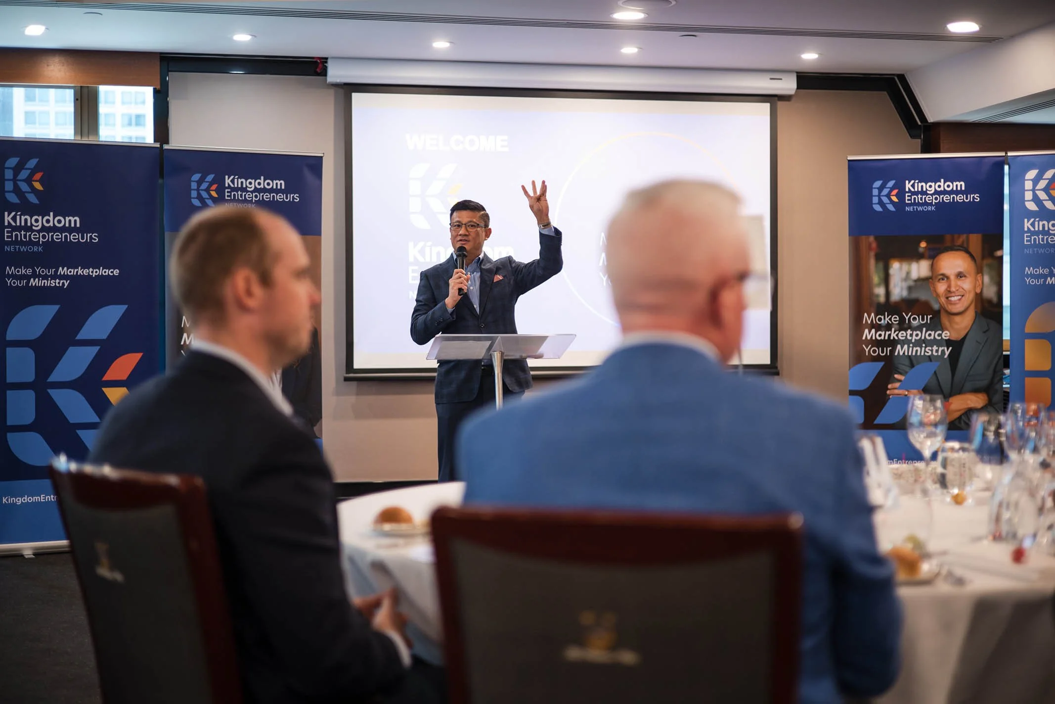 A speaker giving a presentation at a Kingdom Entrepreneurs Network event, with audience members seated at tables, banners displaying the organization's logo and slogan in the background.