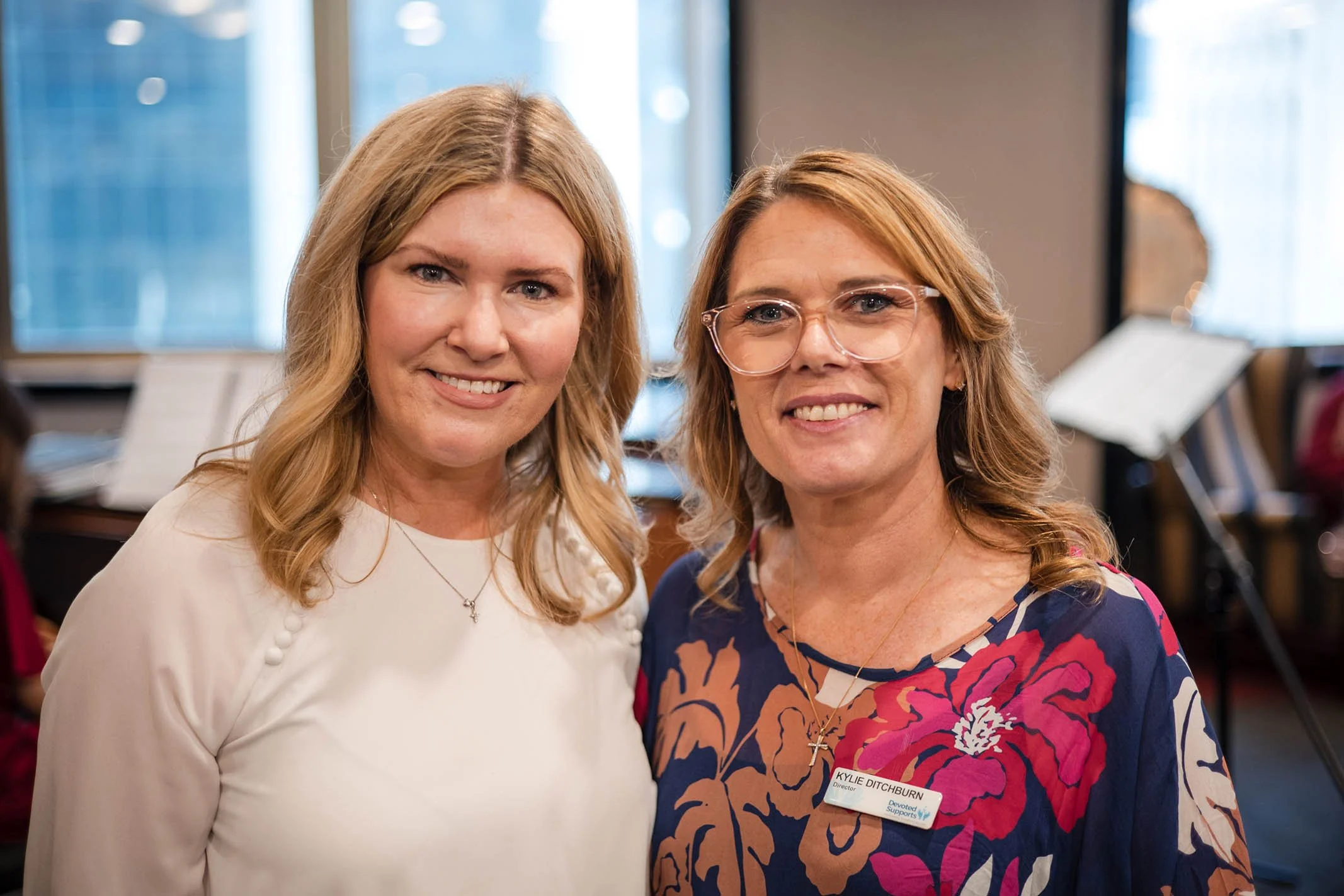 Two women smiling, one wearing a white blouse and the other wearing a floral dress with a name tag that says Kyle Ditchburn, in a room with windows and musical instruments in the background.