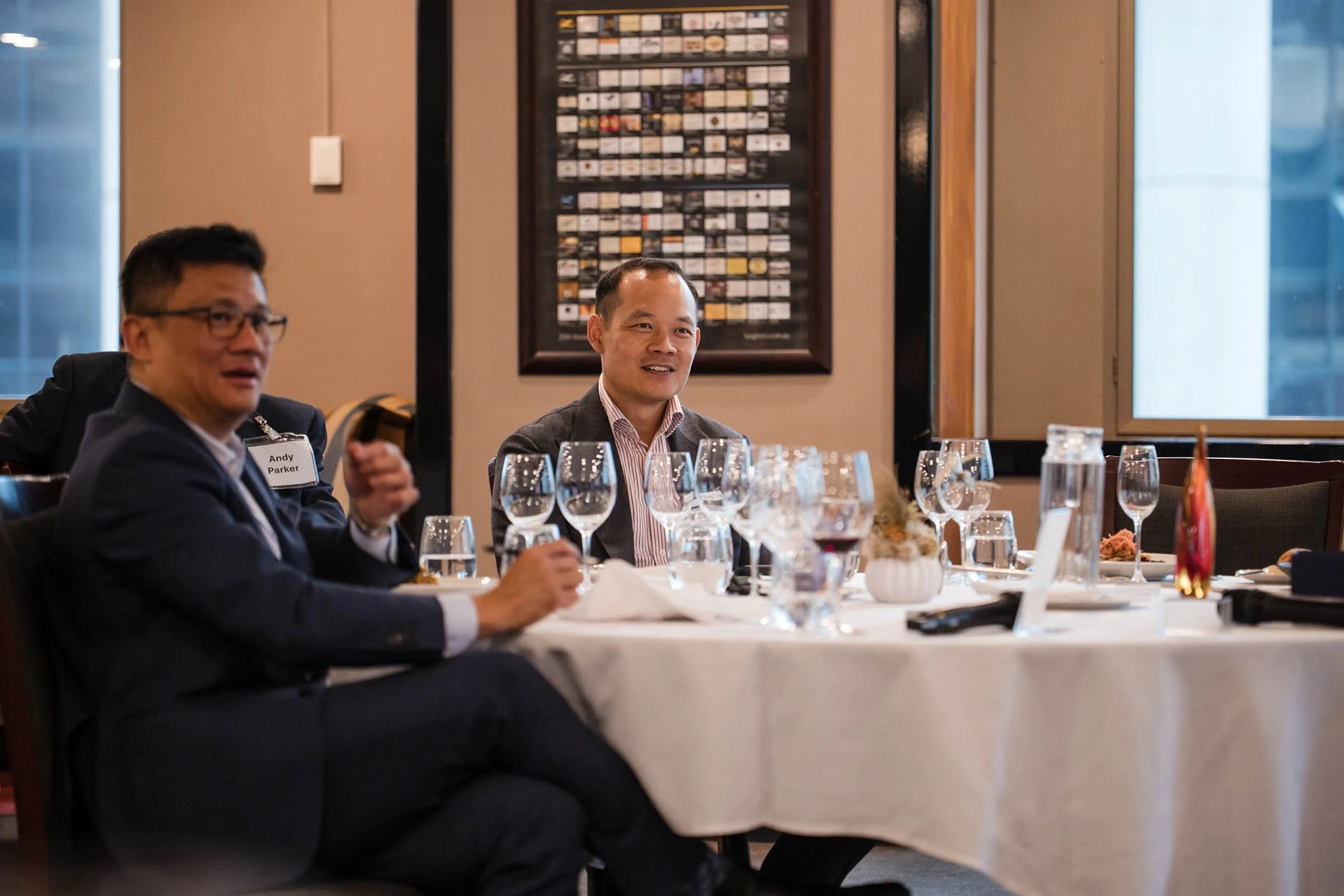 Businessmen seated at a dinner table with glassware, flowers, and decorative items, engaged in conversation at a formal event in a well-lit room with window views.