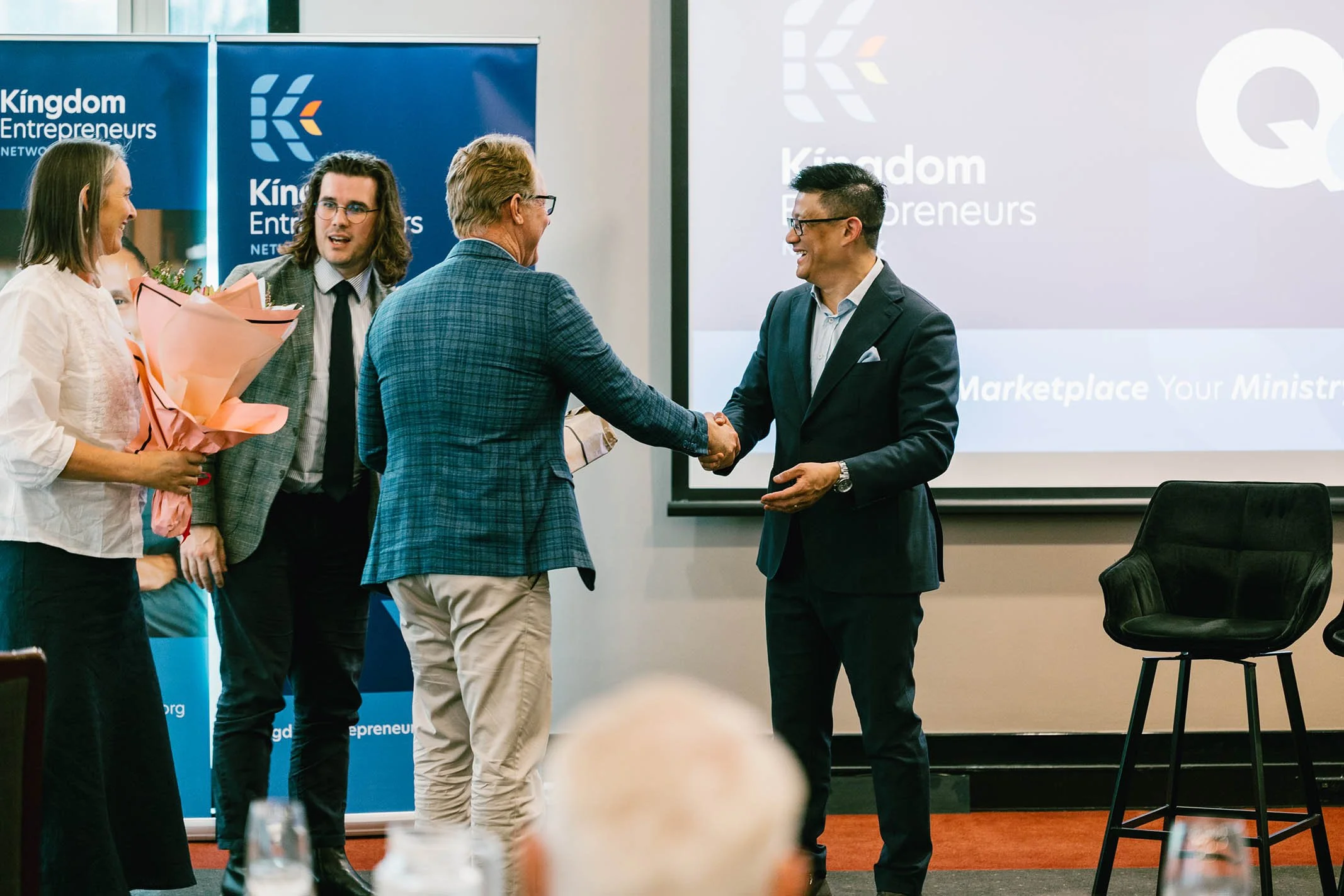 A group of five people at a professional event, with two men shaking hands and a woman holding a bouquet of flowers. There are banners and a projection screen in the background.