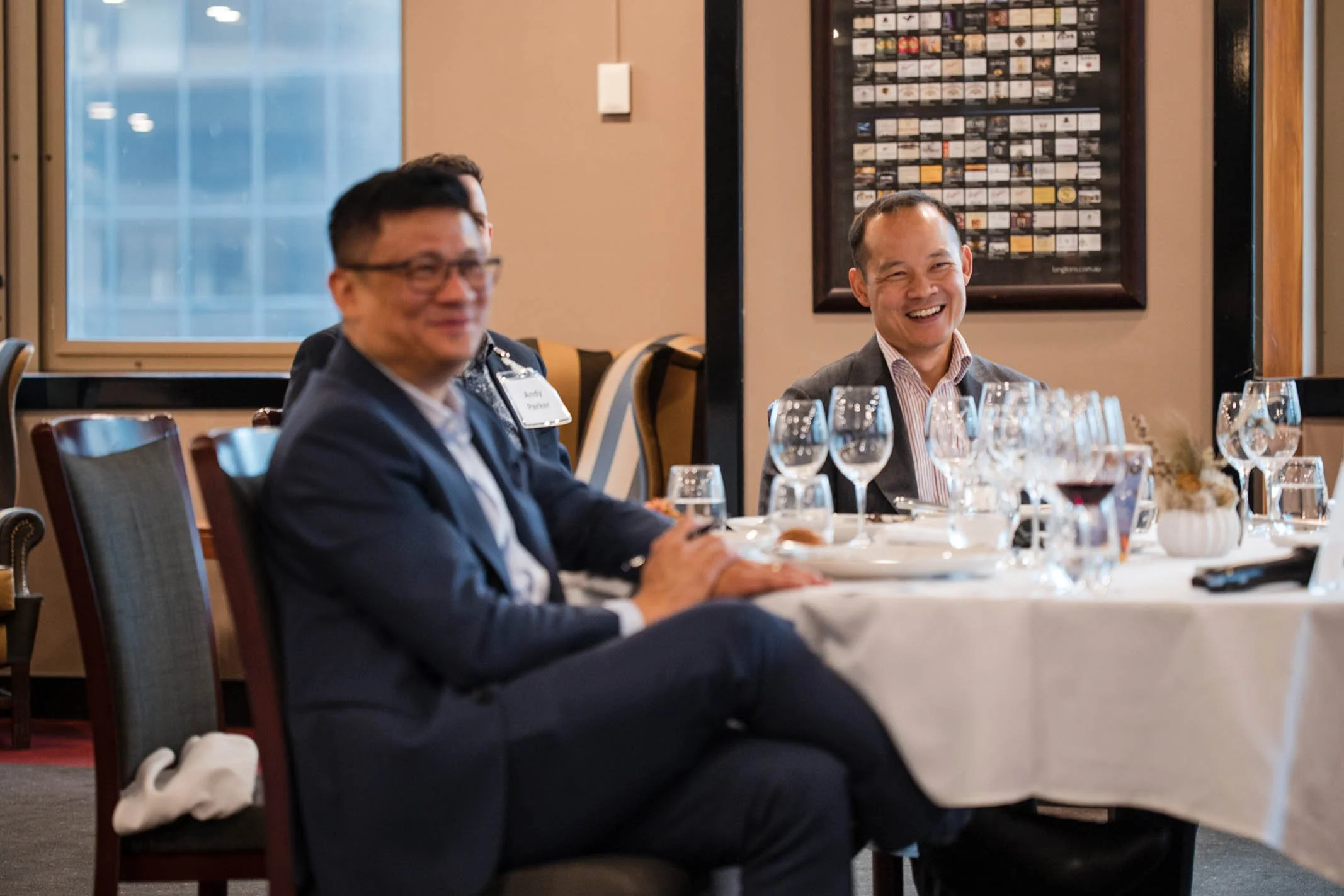 Two men sitting at a dining table, smiling, with wine glasses and a floral centerpiece, in a restaurant or banquet setting.