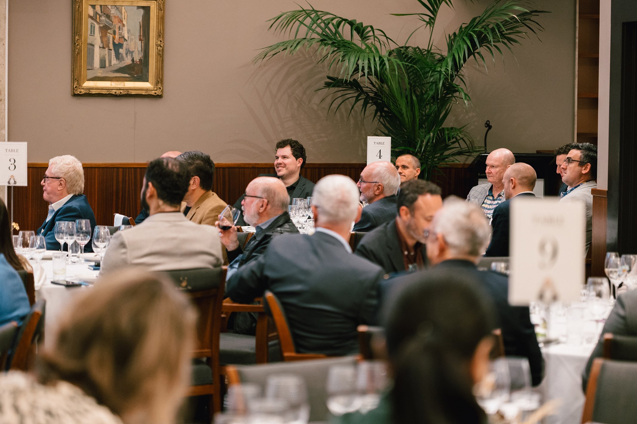 A group of people attending a formal event or dinner, seated at tables with wine glasses, in a room with neutral walls, wood paneling, and a large green plant