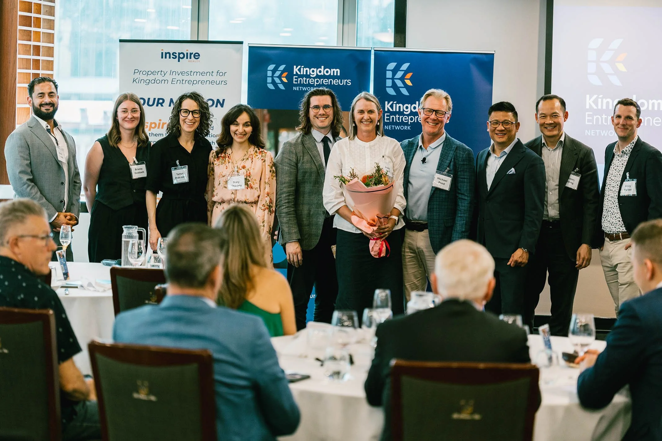 Group of people at a professional event posing for a photo with banners that say 'Kingdom Entrepreneurs' and 'Inspire Realty' in the background, some seated at tables with glasses of water.