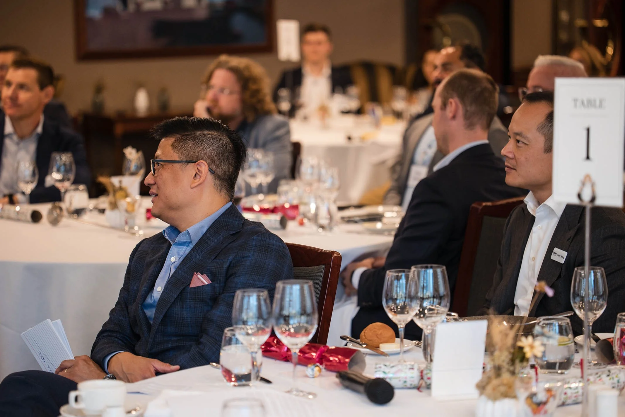 People attending a formal event or conference, seated at round tables with table settings including glasses, plates, and flowers.