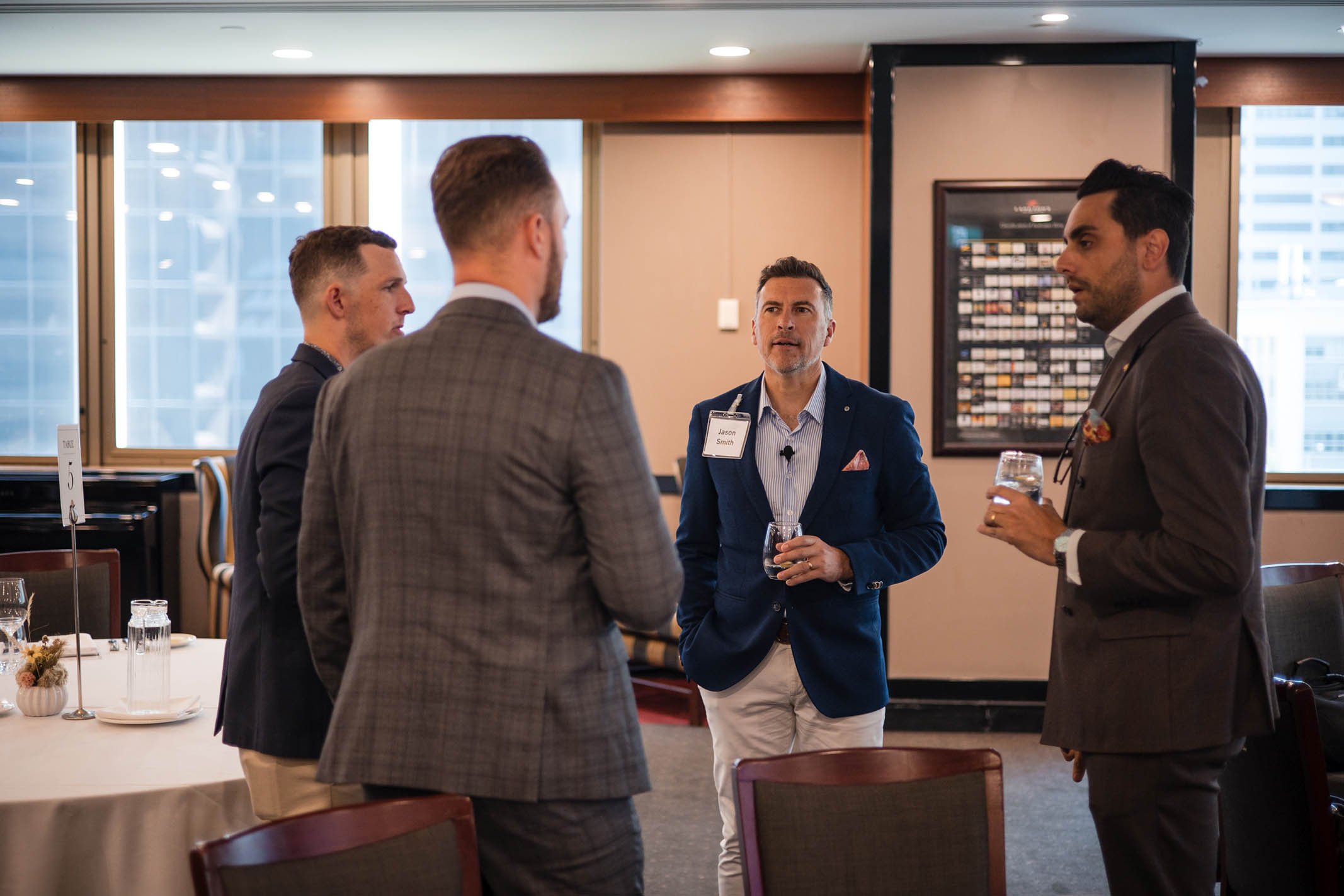 Four men in business attire having a conversation in a conference room, two of them holding glasses of water, with tables and large windows in the background.