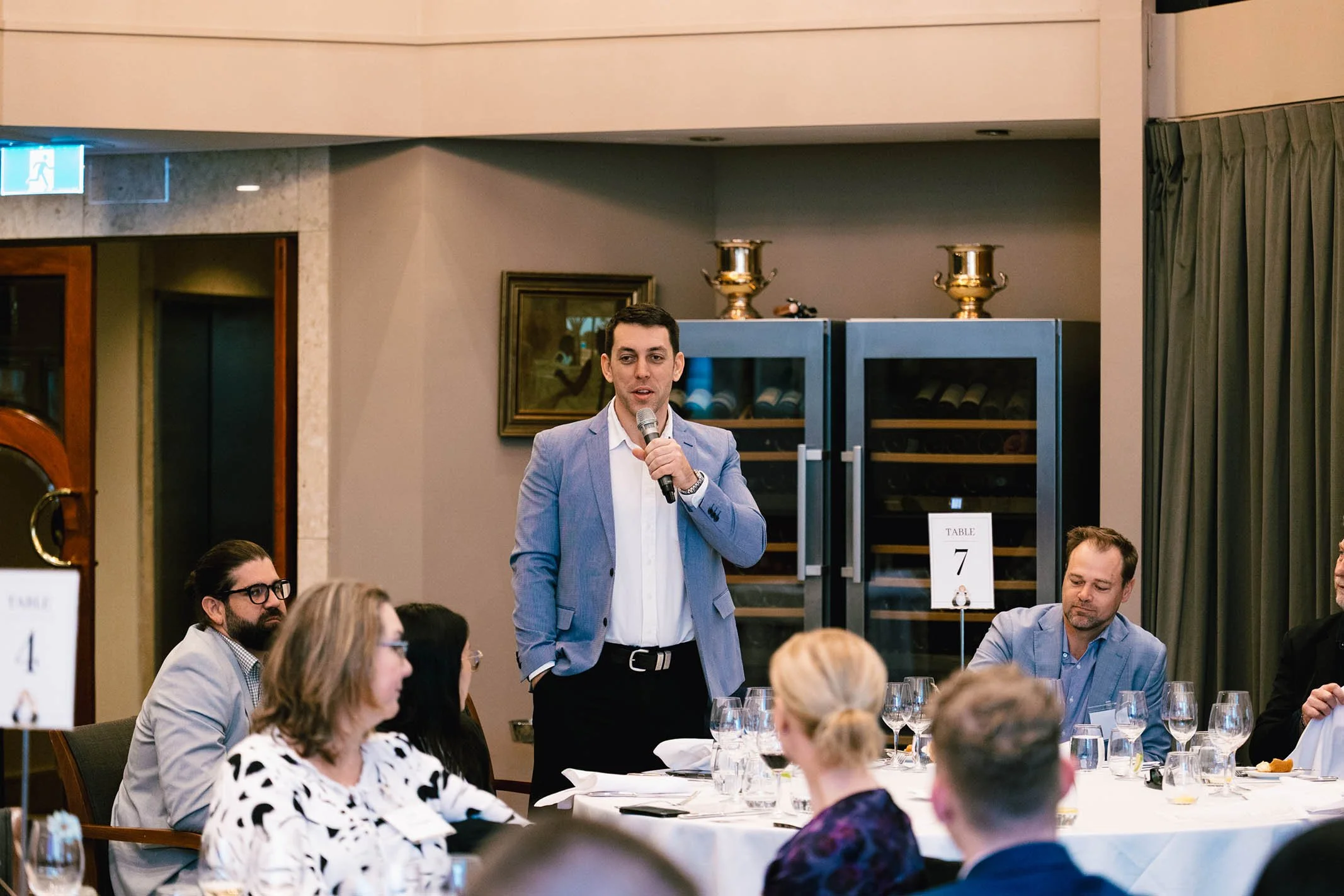 A man in a light blue blazer stands at a round table, speaking into a microphone during a formal event. Several seated attendees, including men and women, listen and look on. The setting appears to be a restaurant or banquet hall with wine cabinets a