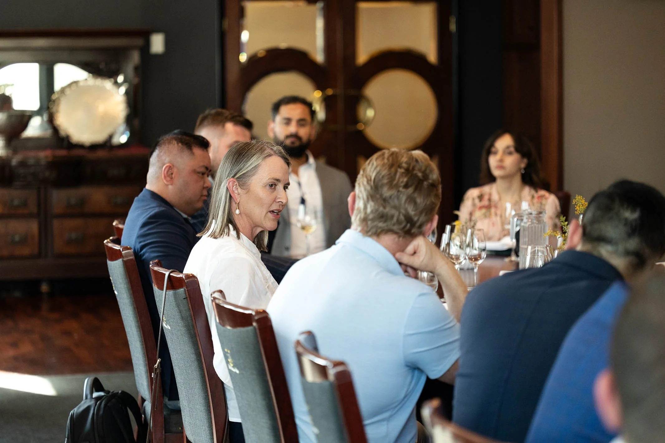 A group of people seated around a conference table engaged in discussion, with some taking notes and others listening attentively, in a formal meeting room.