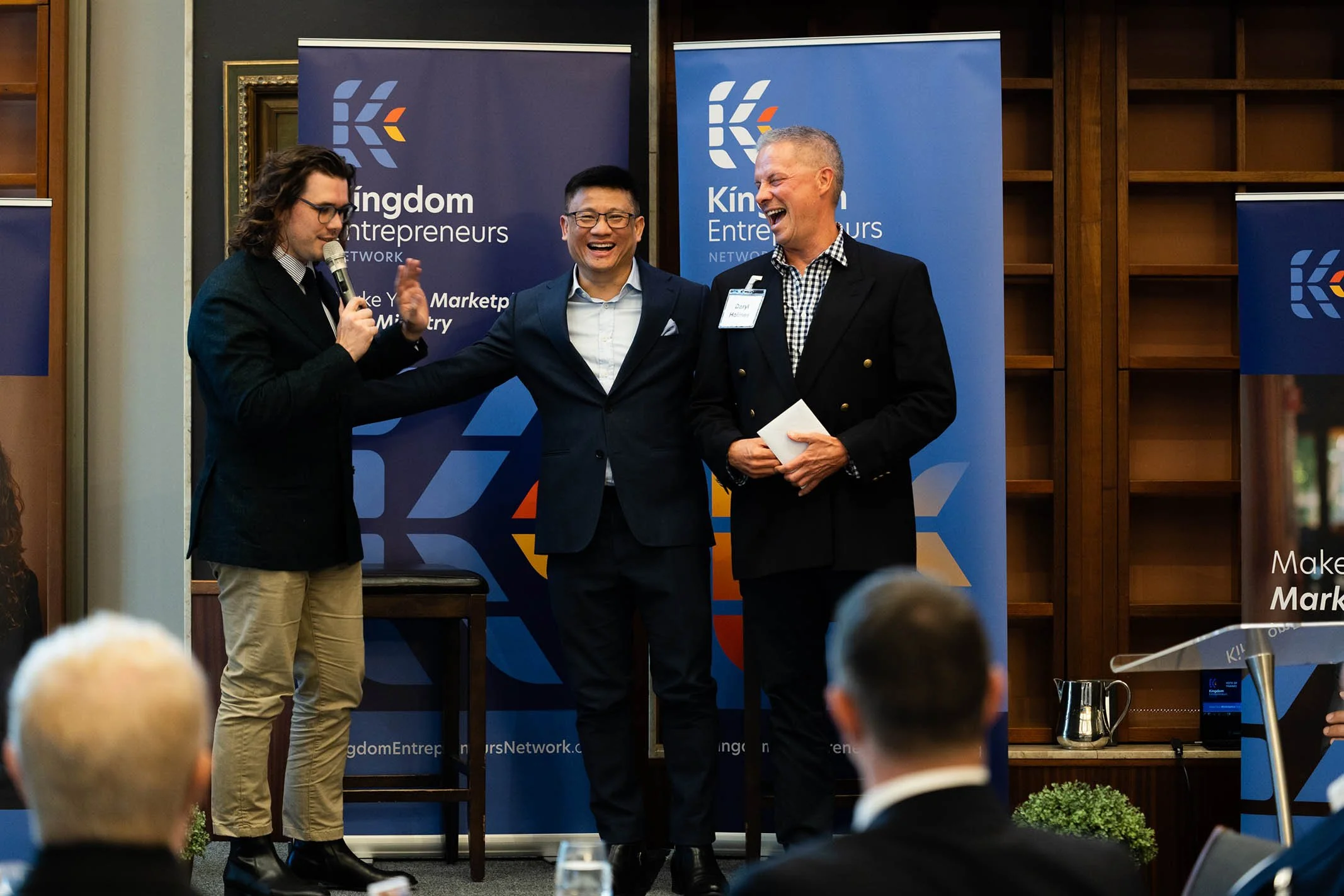 Three men on stage at a networking event, smiling and laughing. One man is speaking into a microphone, another man has his arm around a third man. Behind them are blue banners that read "Kingdom Entrepreneurs Network."