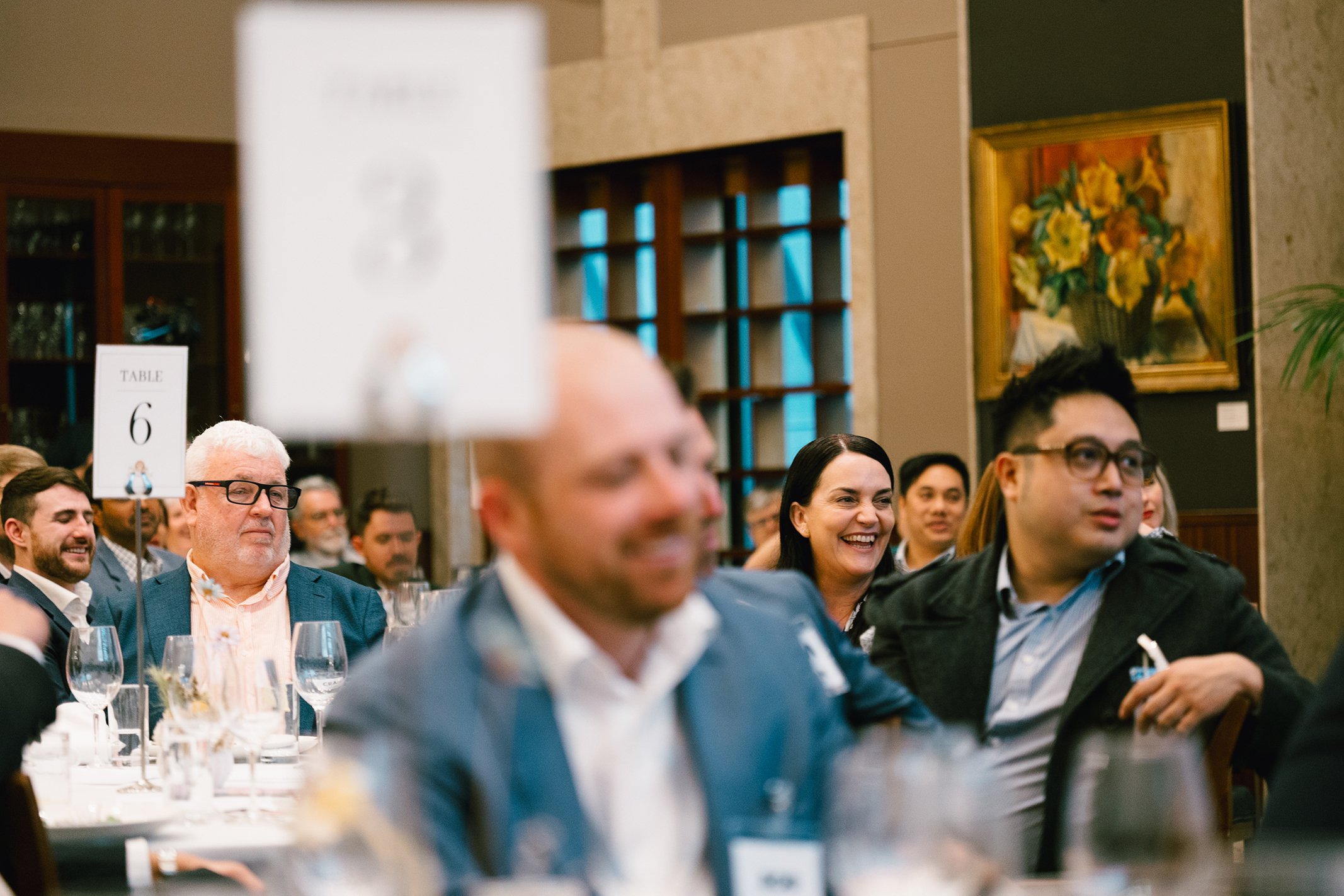 A group of people sitting at a banquet table in a well-lit room, some smiling and laughing. The focus is on a woman in the center smiling, while other guests are engaged and attentive. There is a sign indicating 'Table 6' on the table.