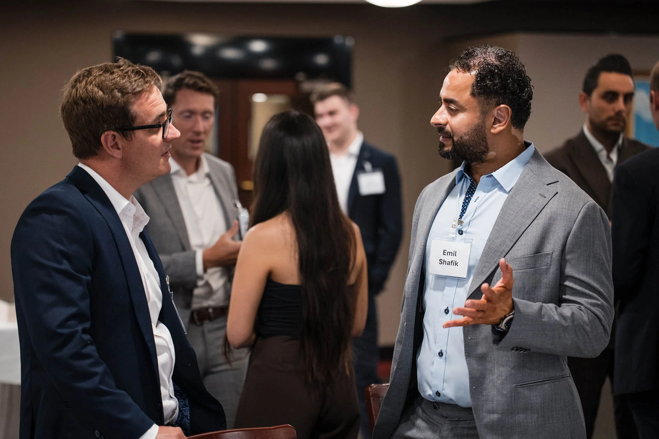 Man with a gray suit and a name tag labeled Emil Shafik talking to a young man in a navy suit at a professional networking event, with other attendees in the background.