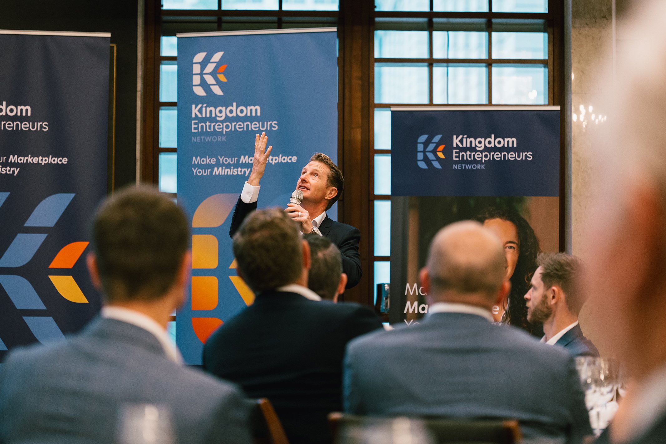 A man in a black suit is giving a presentation at a conference, holding a microphone in one hand and gesturing upward with the other. Several attendees are seated in front of him. The background features banners for 'Kingdom Entrepreneurs Network' wi