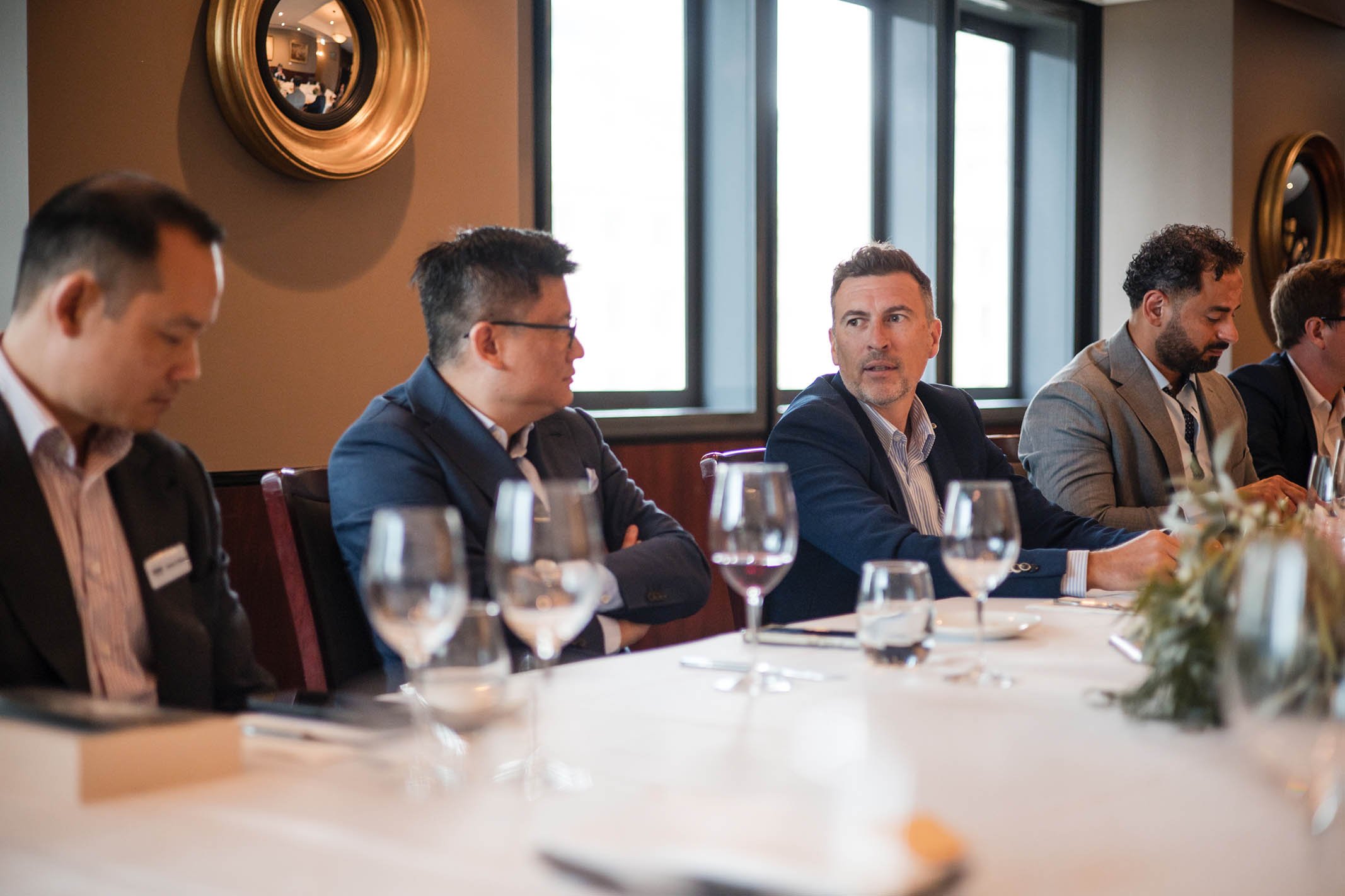 Business professionals in formal attire attending a meeting or conference at a long table with glassware, in a well-lit room with large windows and decorative wall mirrors.