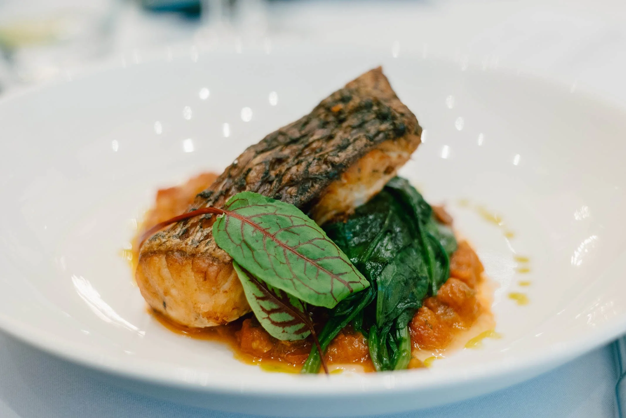 A plated dish featuring a grilled salmon fillet with crispy skin, served with wilted greens on a bed of orange-colored sauce.