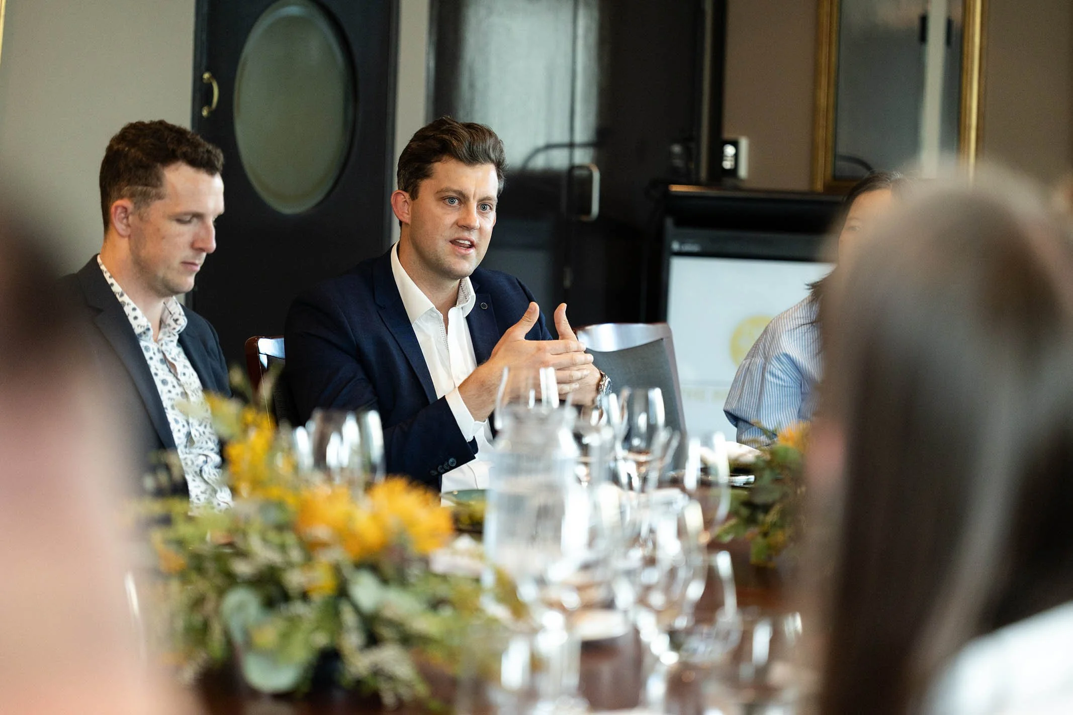 A man in a dark suit and white shirt speaking at a meeting, with other people seated at the table, during a formal gathering or discussion.