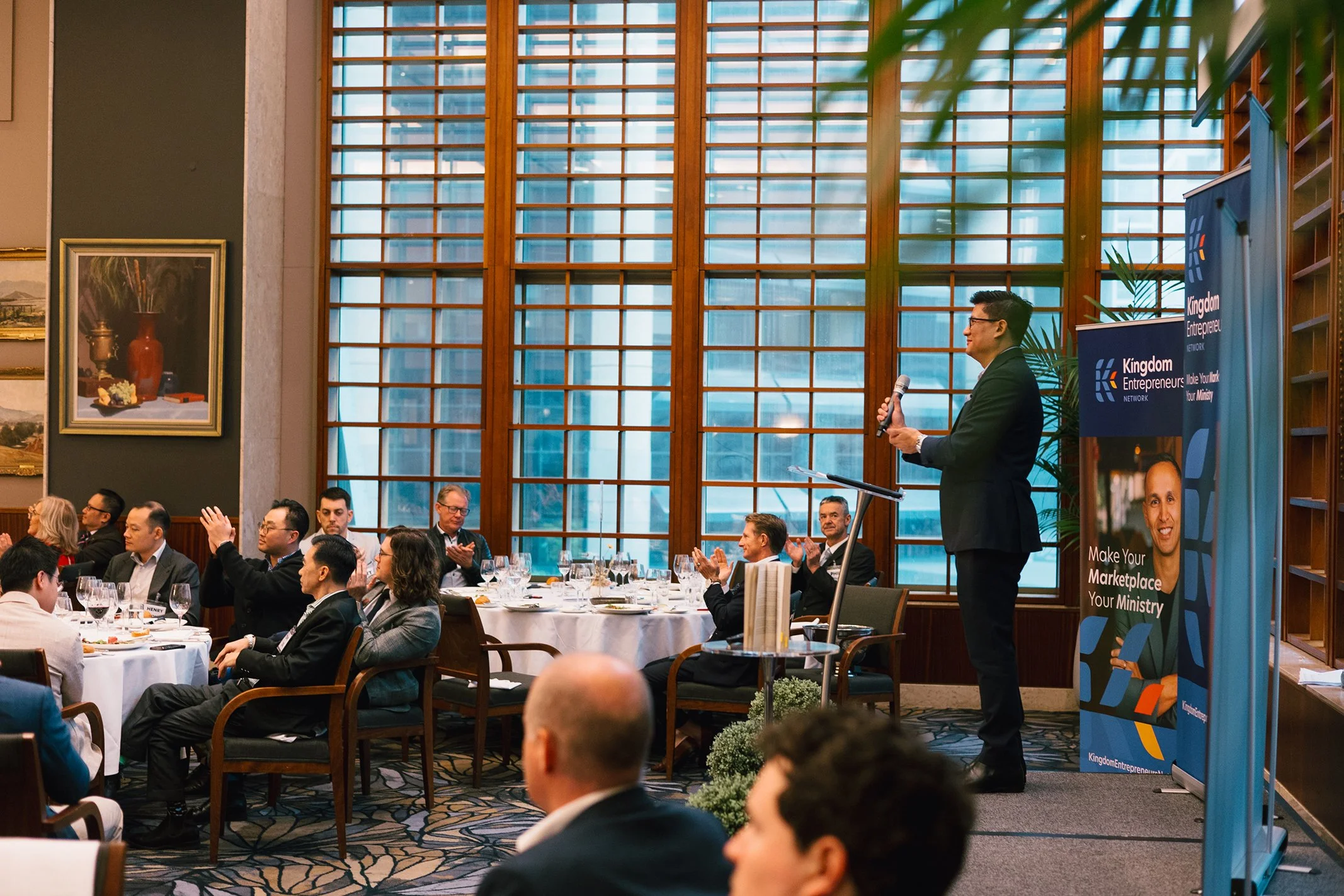 A man in a suit speaking into a microphone at a conference or business event, standing beside banners for Kingdom Entrepreneurs, with attendees seated at round tables applauding.
