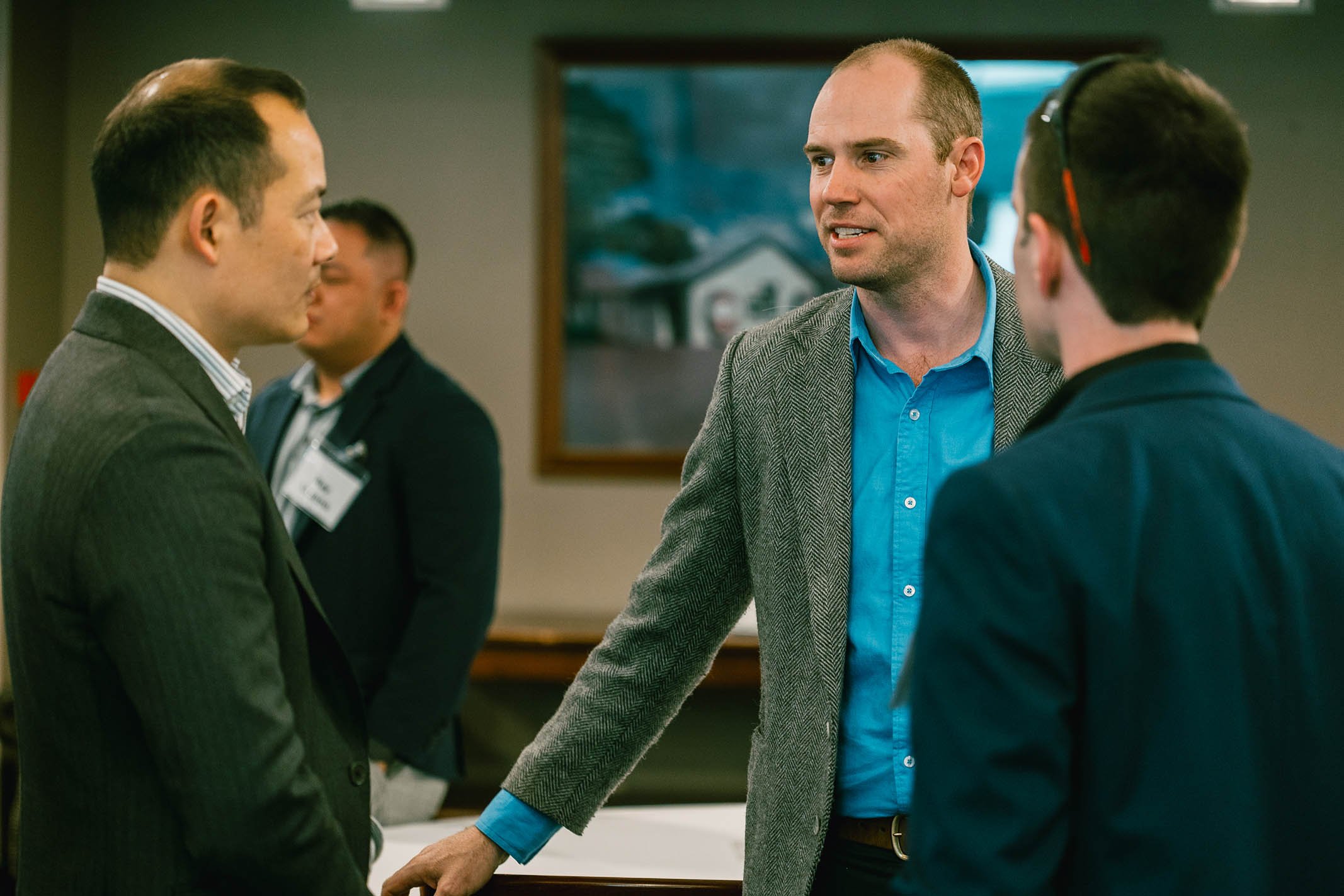 Four men engaged in conversation in a professional setting. One man with a shaved head and gray blazer is speaking to a man with dark hair and a man bun, dressed in a dark blazer. Two other men are in the background, one with a badge, all wearing bus